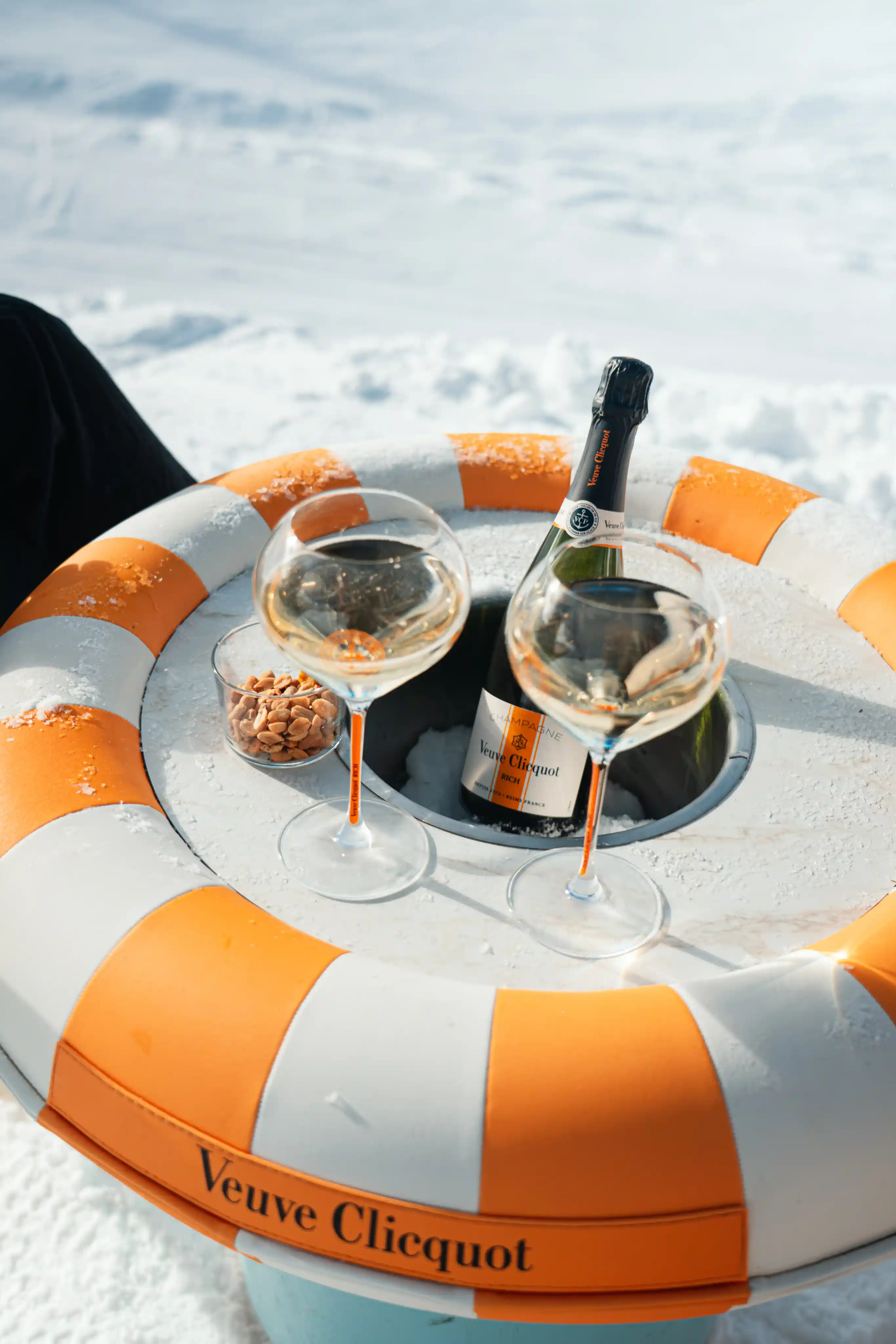 Bouteille de champagne Veuve Clicquot placée dans un seau à glace intégré à une bouée orange et blanche sur la neige, avec deux coupes de champagne remplies et un petit bol de cacahuètes.