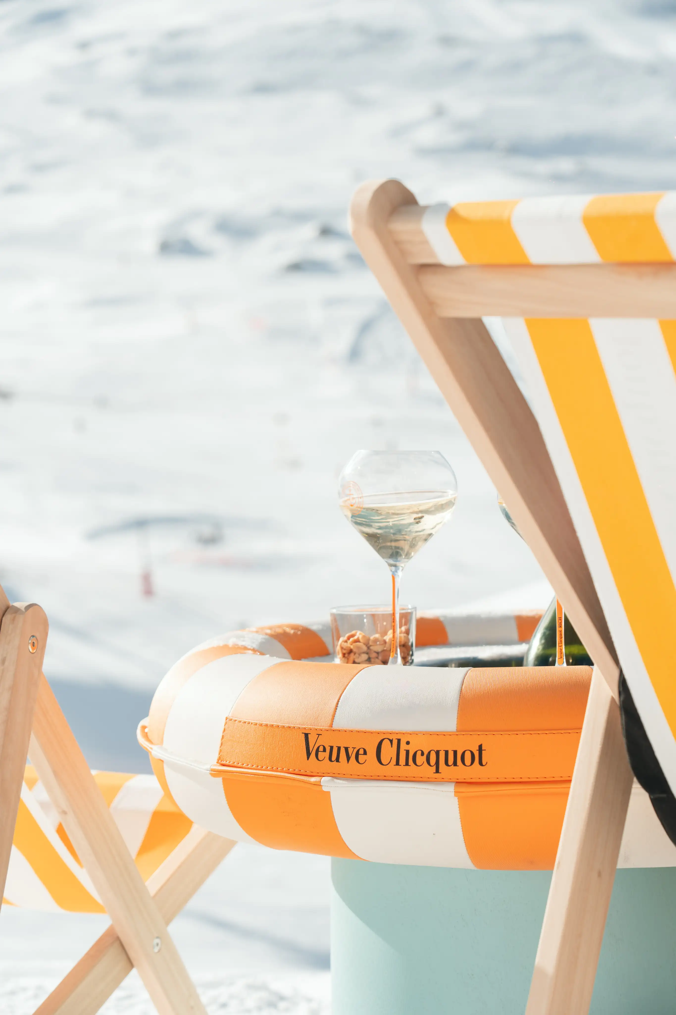 Chaise longue rayée jaune et blanche près d'une bouée Veuve Clicquot contenant un verre de champagne et un petit bol de noix, avec un décor enneigé en arrière-plan.