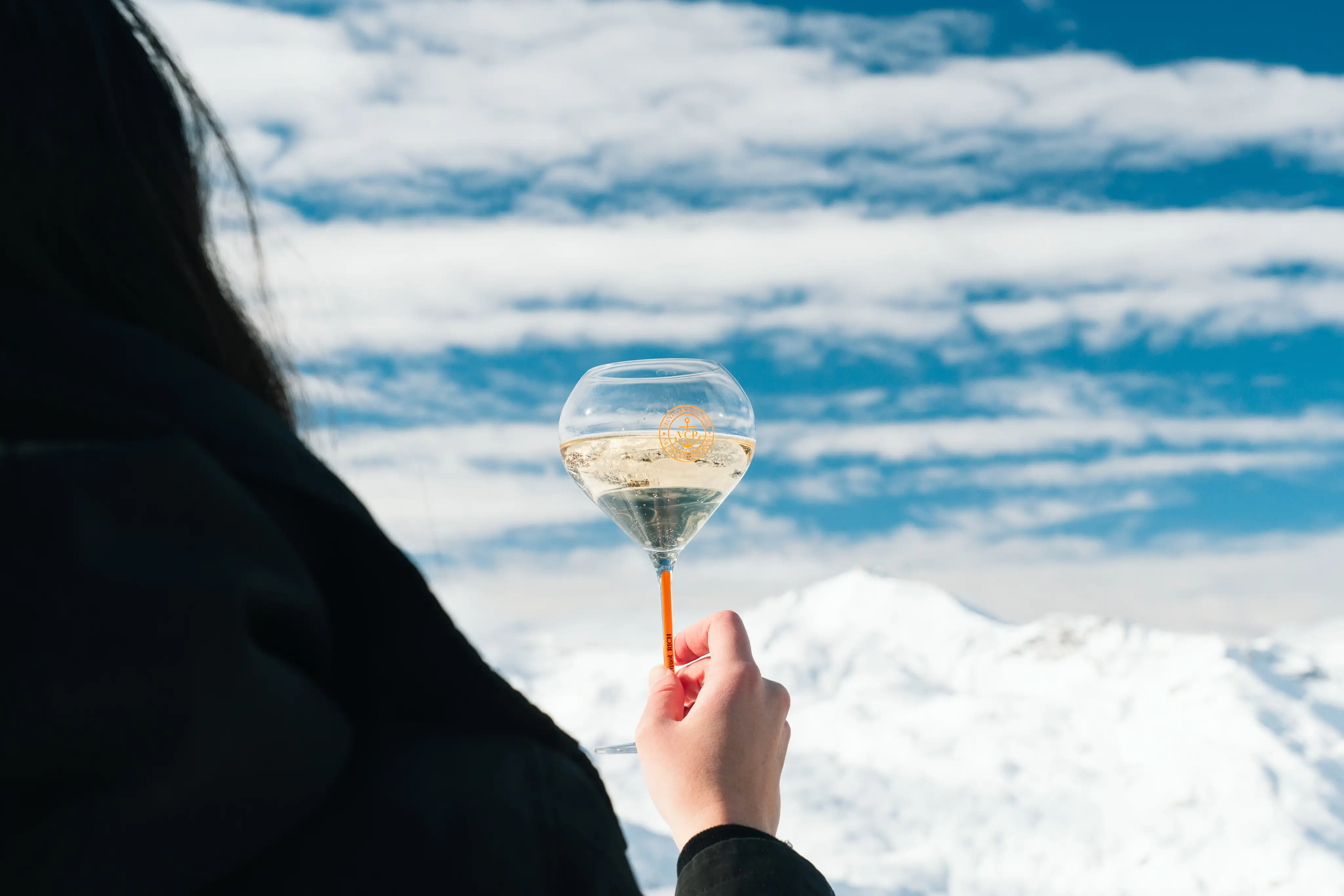 Person vue de dos tenant un verre à pied de champagne transparent rempli de vin mousseux clair, avec un ciel bleu nuageux et des montagnes enneigées en arrière-plan.