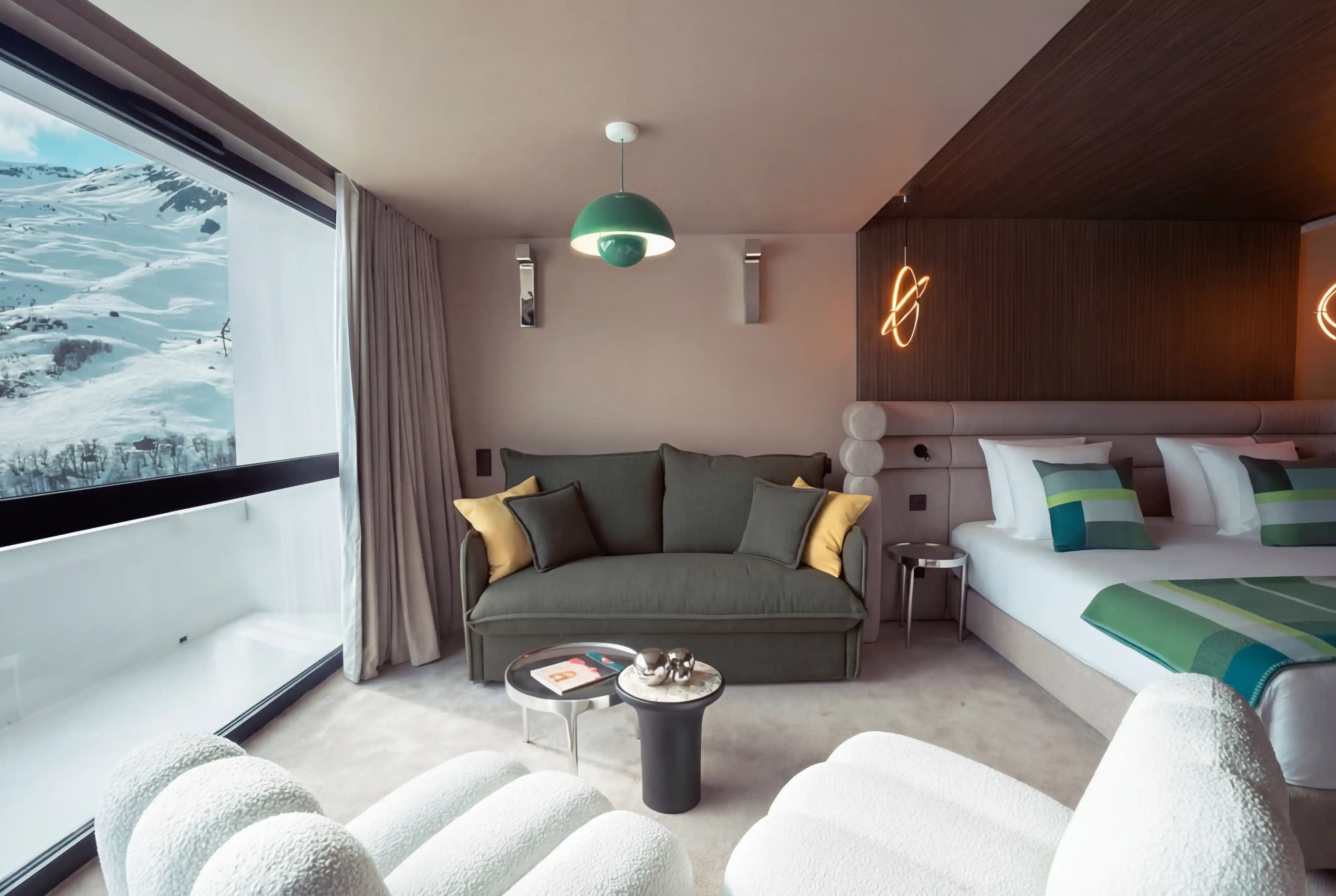 Chambre d'hôtel moderne avec un grand lit double équipé de coussins verts, un canapé vert avec coussins jaunes, deux petites tables au centre, deux fauteuils blancs en premier plan, une grande baie vitrée donnant sur un paysage enneigé de montagnes, et un éclairage suspendu et mural décoratif.