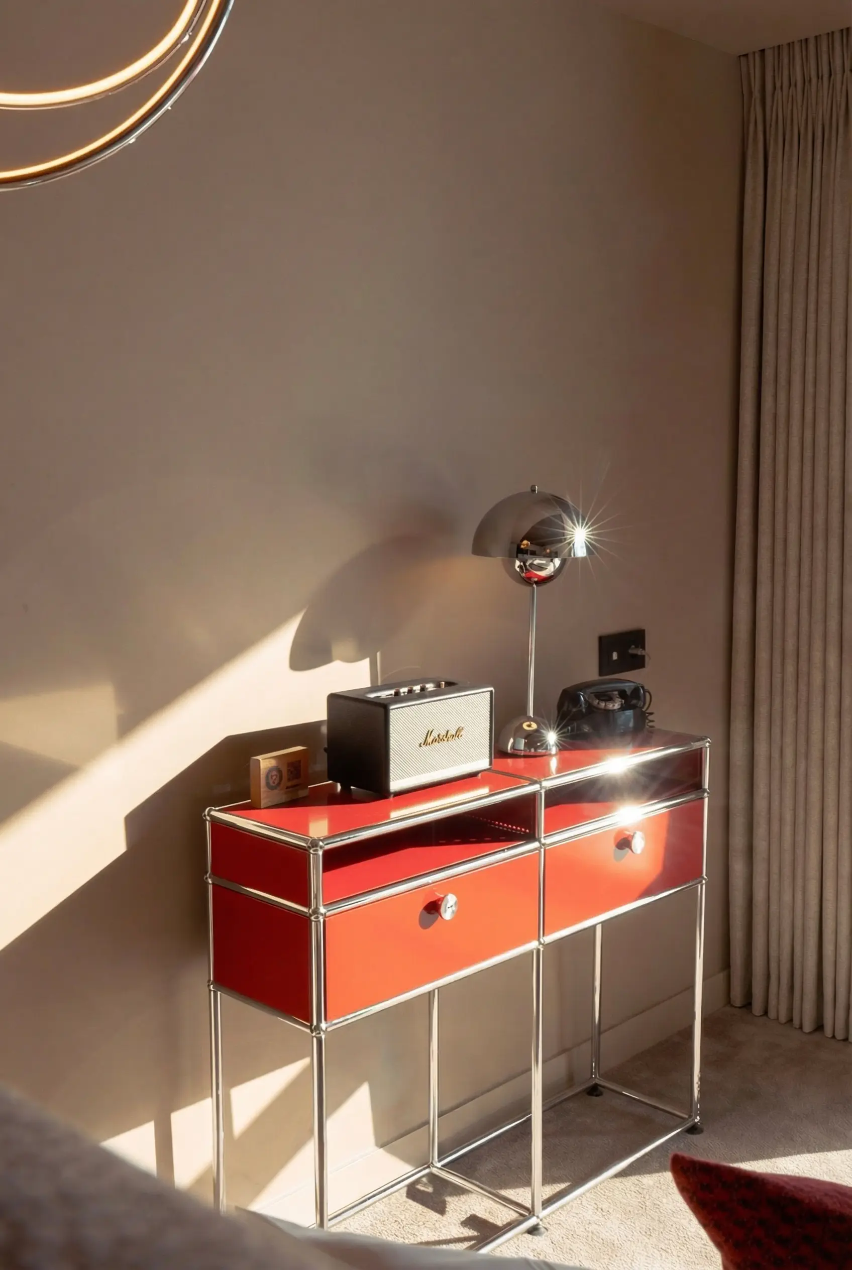 Console moderne à deux tiroirs rouges avec une lampe en métal chromé, un haut-parleur Marshall et un téléphone vintage noir posés dessus, éclairée par la lumière du jour près d'un mur beige et de rideaux beiges.