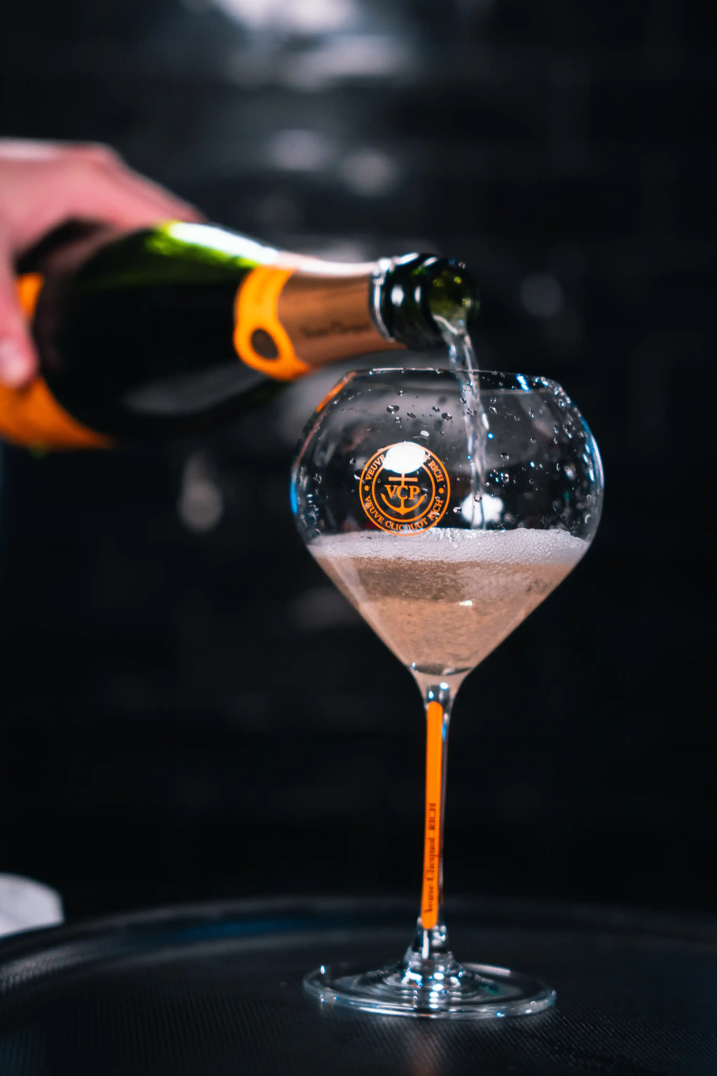 Une bouteille de champagne Veuve Clicquot est versée dans un verre à pied marqué du logo VCP, avec du liquide pétillant et des gouttes sur le verre.