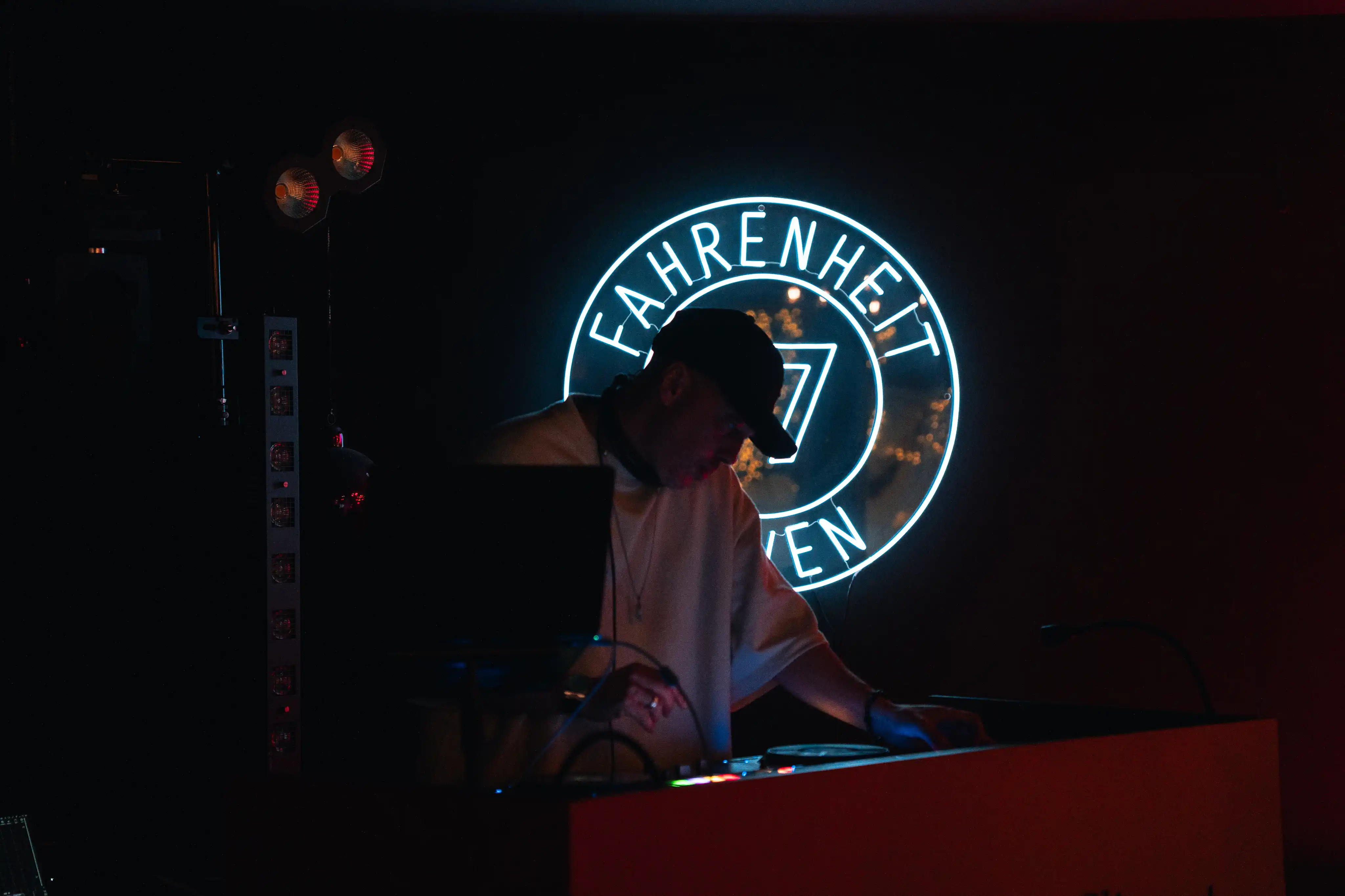 Un DJ portant une casquette noire et un t-shirt clair est en train de mixer de la musique devant un panneau lumineux circulaire avec l'inscription "FAHRENHEIT SEVEN" dans une pièce sombre.