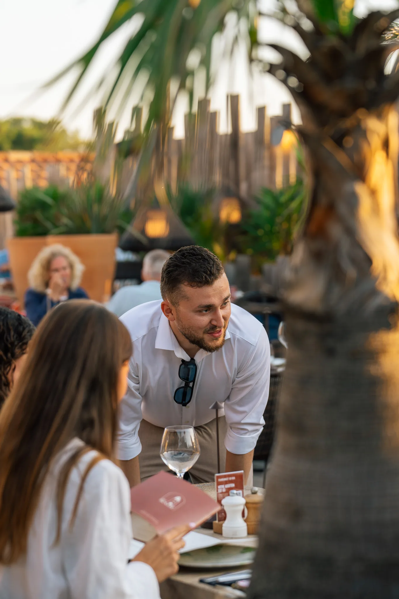 Un serveur souriant en chemise blanche parlant à une cliente tenant un menu dans un restaurant en plein air avec des palmiers et un décor naturel en arrière-plan.