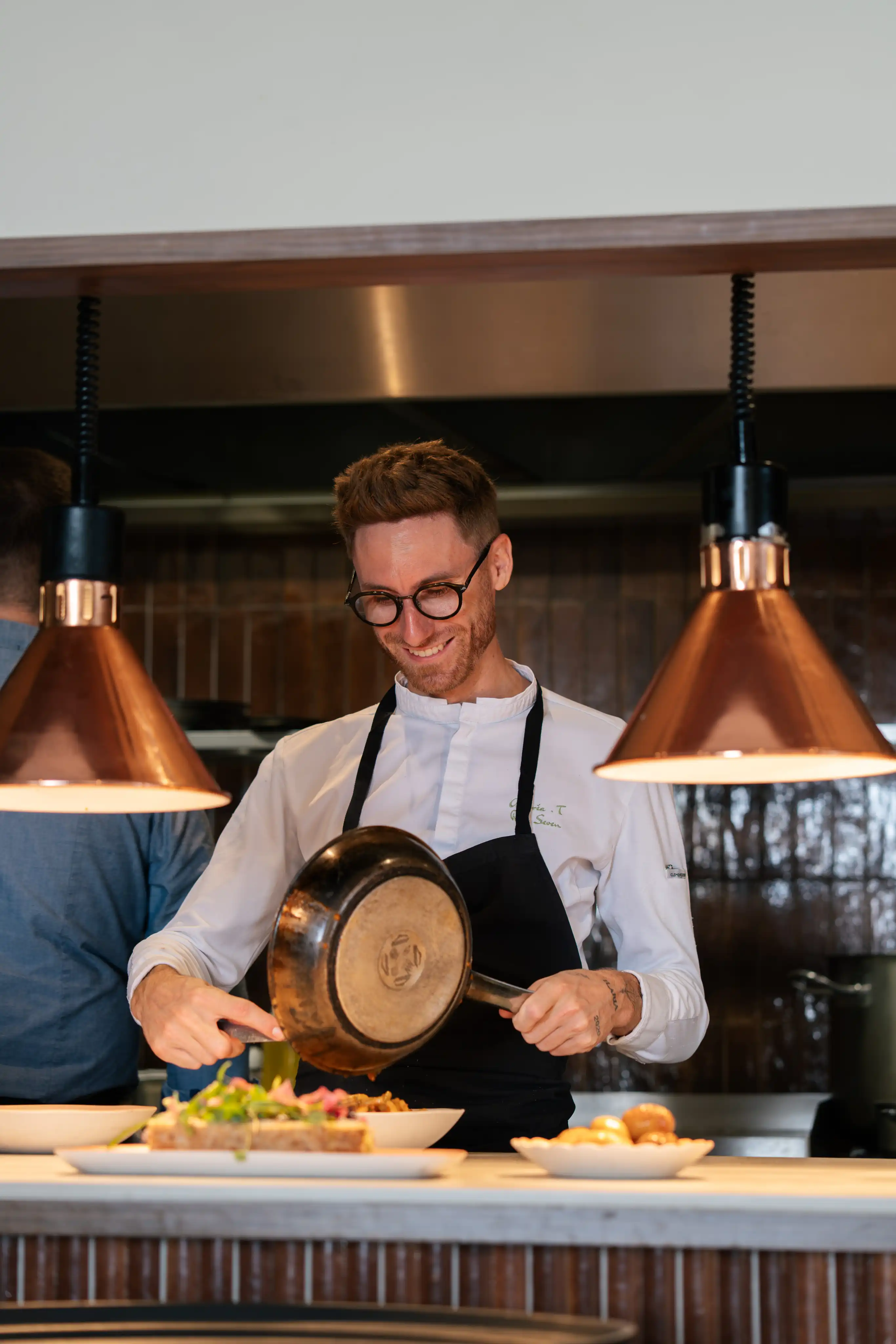 Un chef cuisinier souriant en blouse blanche et tablier noir verse un plat à partir d'une poêle, dans une cuisine professionnelle sous deux lampes suspendues.