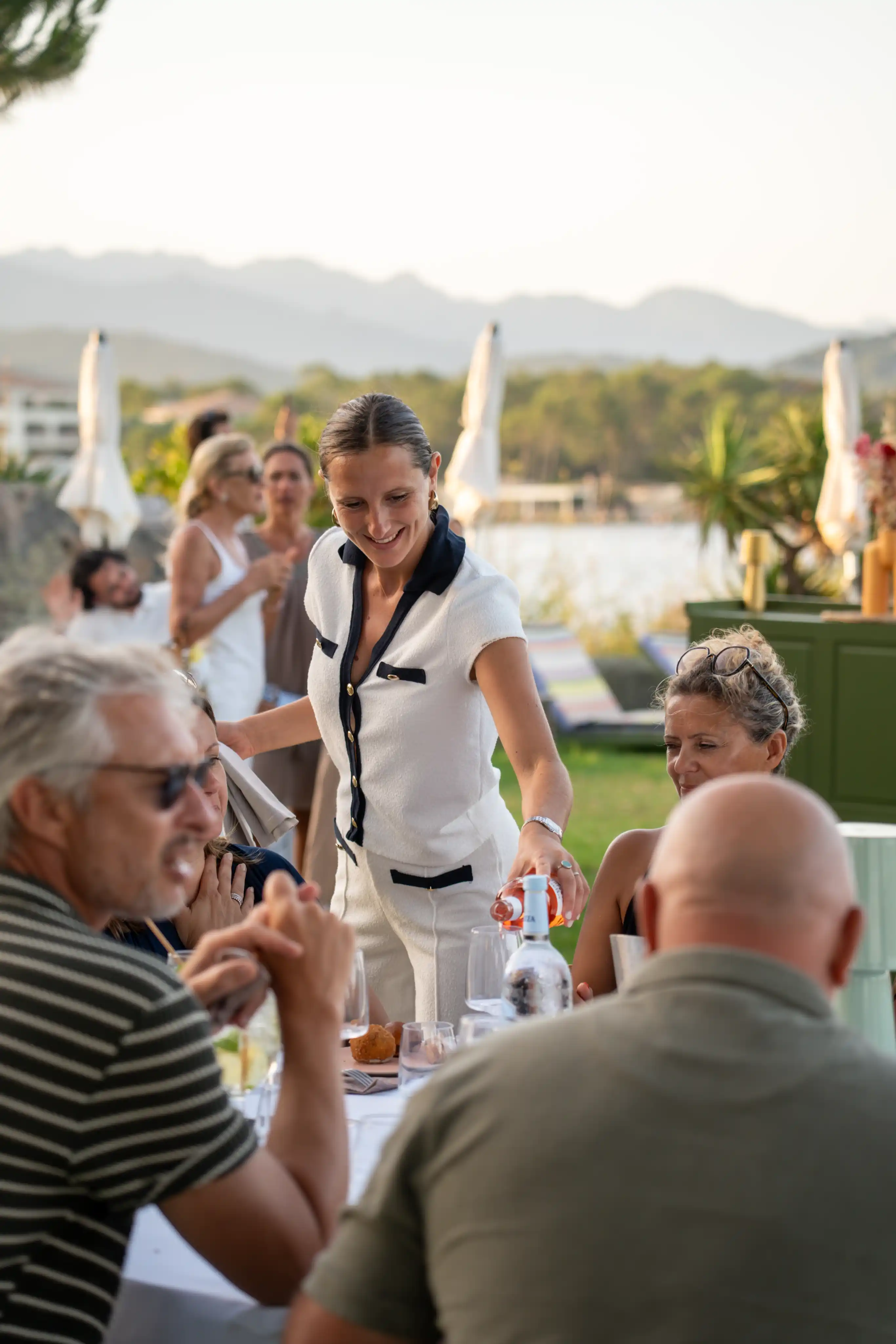 Serveuse souriante servant du vin à une table extérieure où plusieurs personnes adultes discutent, avec un paysage de montagnes, arbres et parasols en arrière-plan.