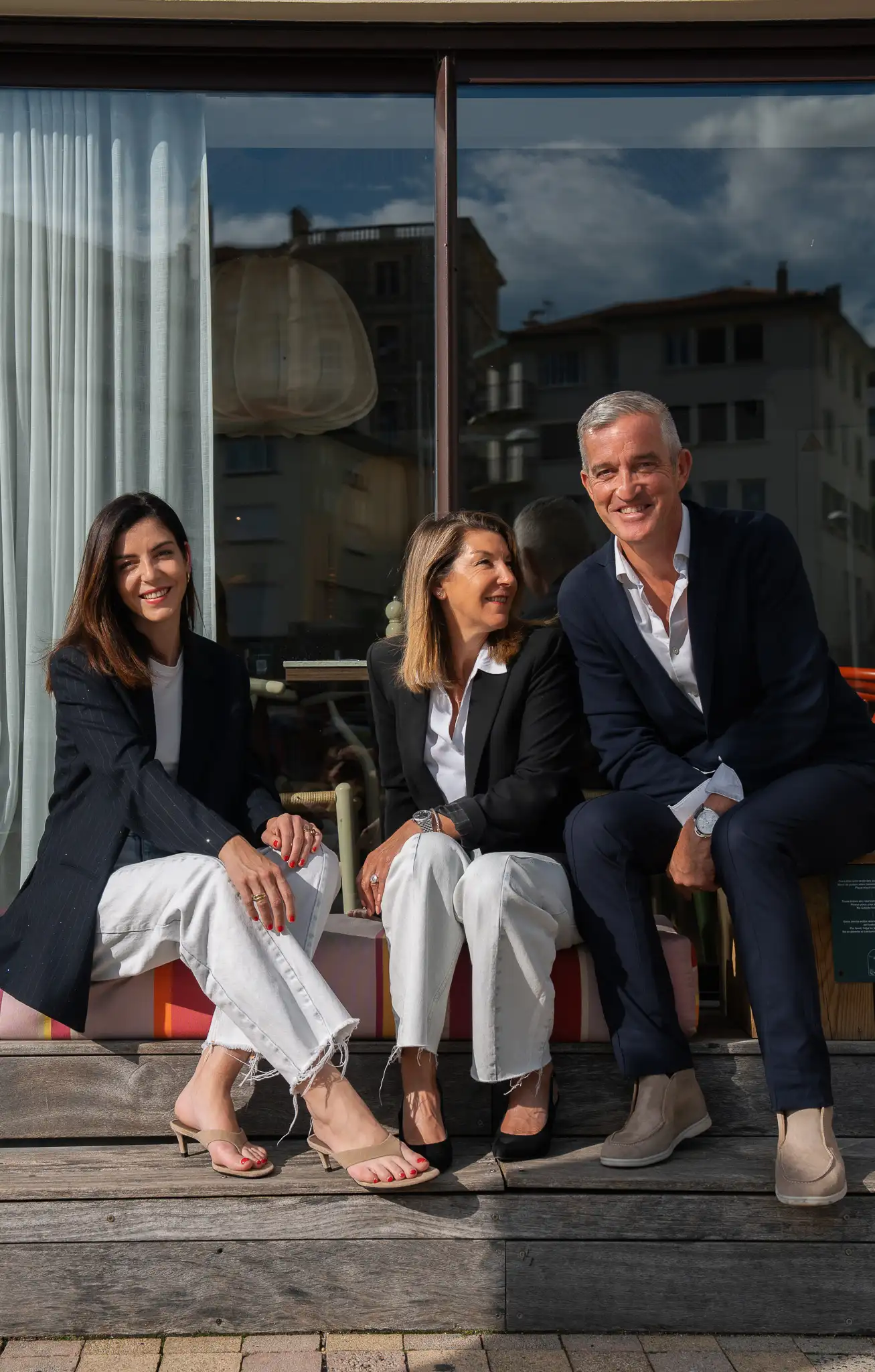 Trois adultes assis sur un banc en bois devant une fenêtre: deux femmes portant des blazers noirs et des pantalons blancs, et un homme en costume bleu avec chemise blanche, tous souriants à la lumière naturelle.