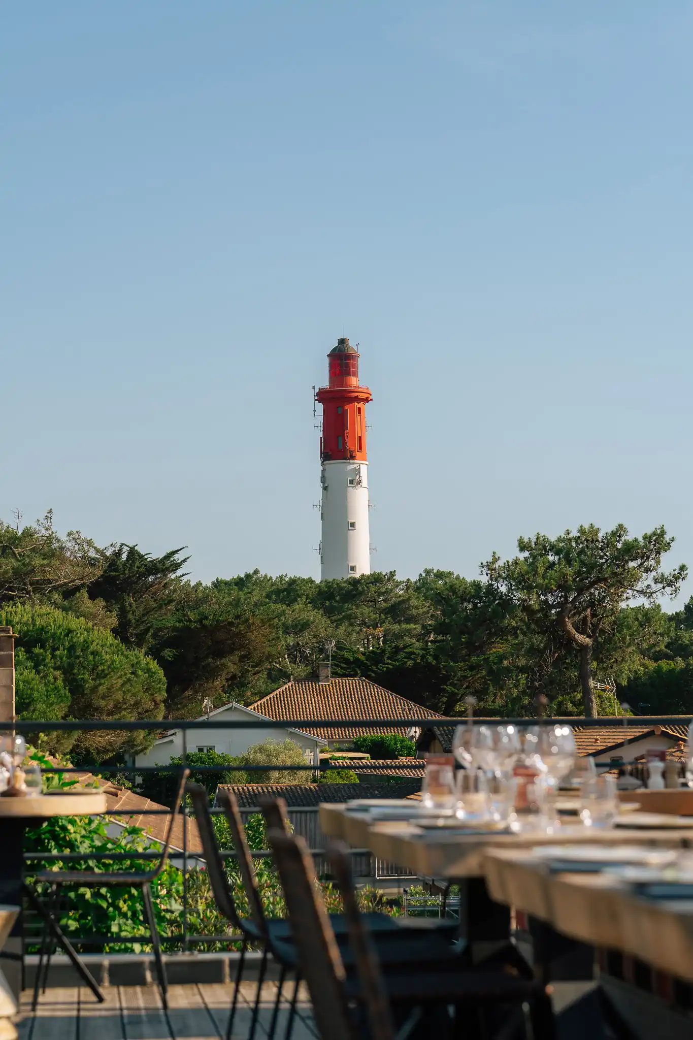 Phare blanc et rouge se détachant derrière des arbres verdoyants et des toits de maisons, avec des tables et chaises de restaurant floues au premier plan.