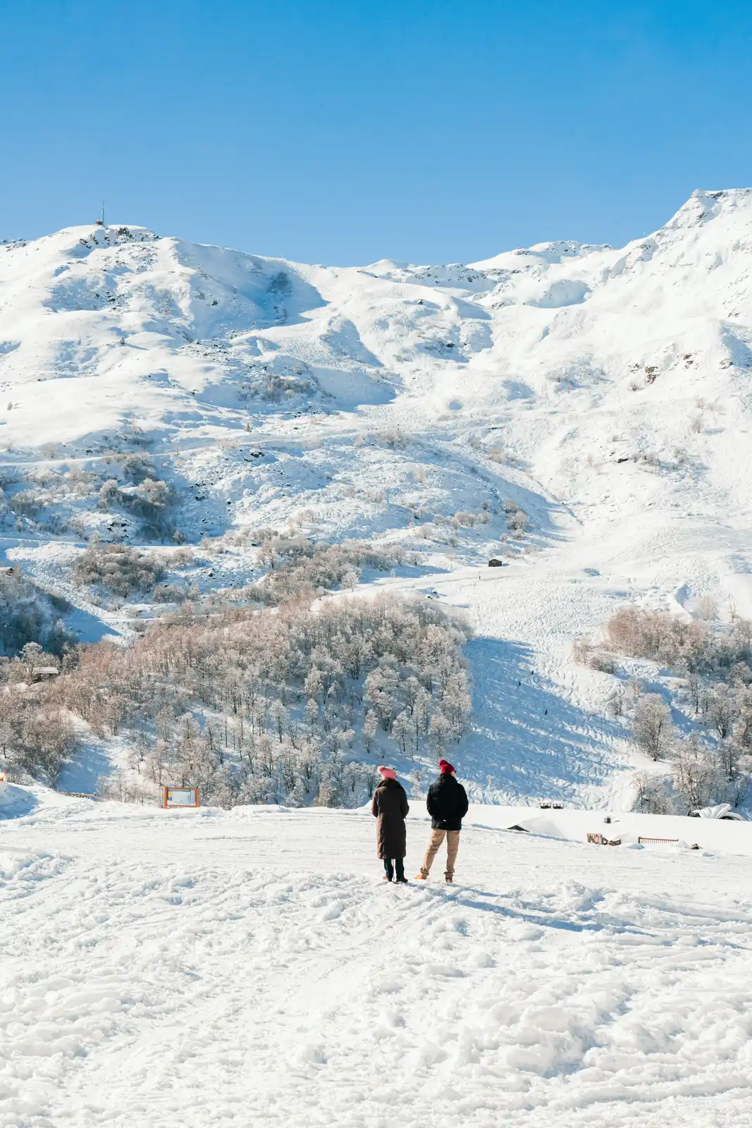 Deux personnes debout dans la neige, regardant des montagnes enneigées et des arbres partiellement couverts de neige sous un ciel bleu clair.