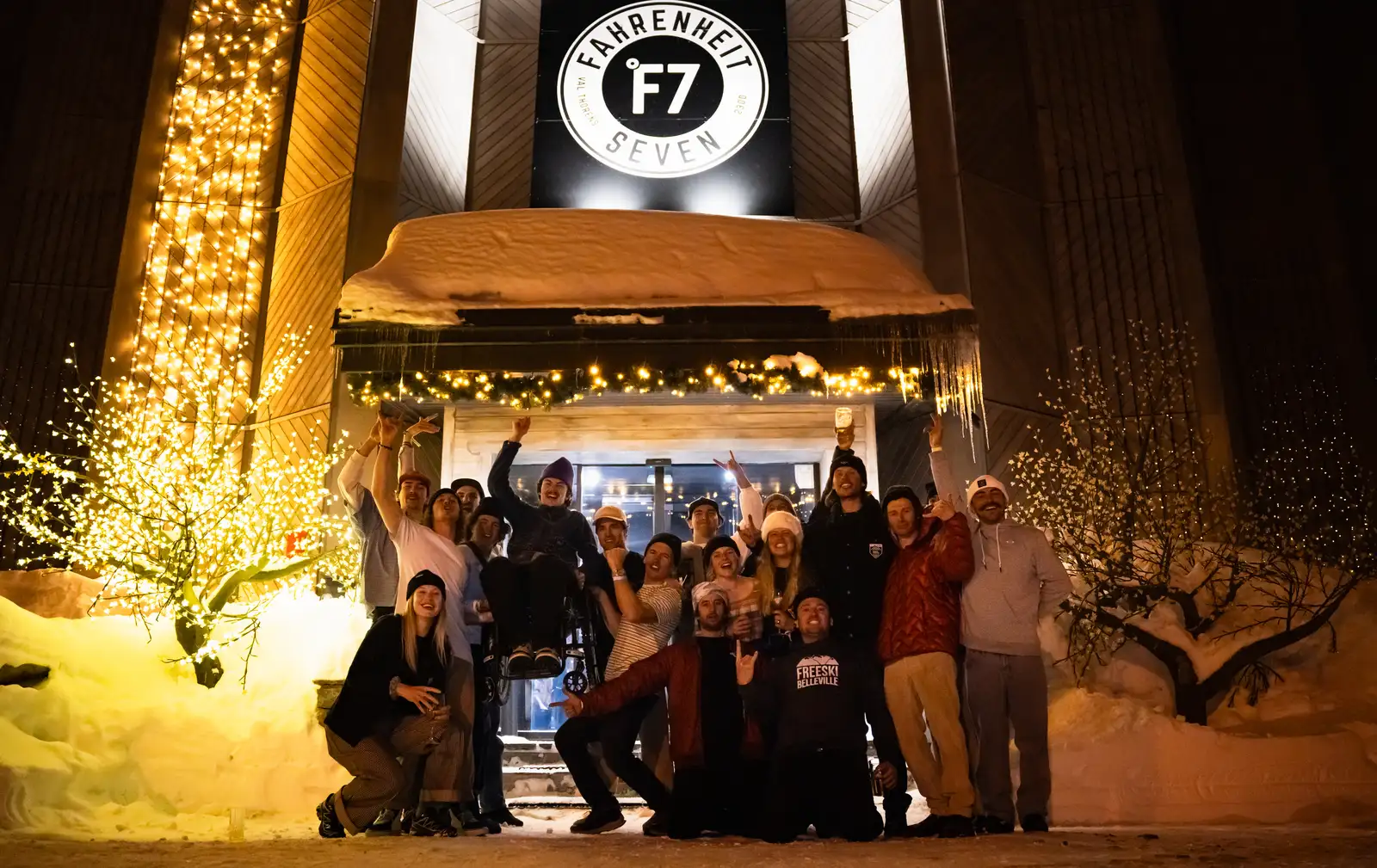 Groupe de personnes joyeuses posant devant un bâtiment décoré pour l'hiver avec des lumières et de la neige, sous un panneau indiquant Fahrenheit 7.