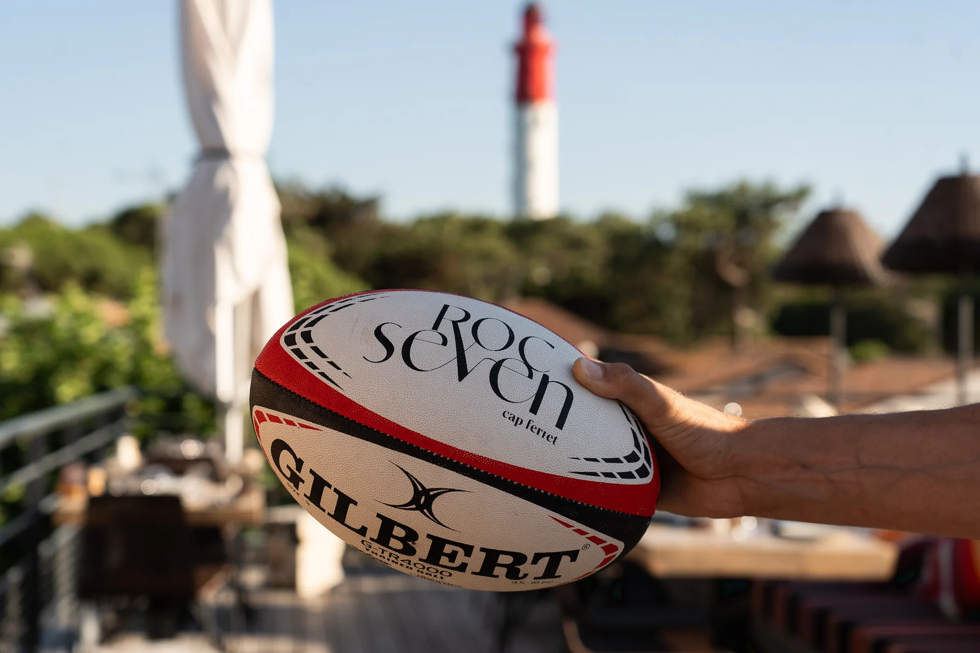 Une main tenant un ballon de rugby blanc, rouge et noir marqué Gilbert et Roc Seven cap ferret, avec un arrière-plan flou comprenant un phare rouge et blanc et des parasols.