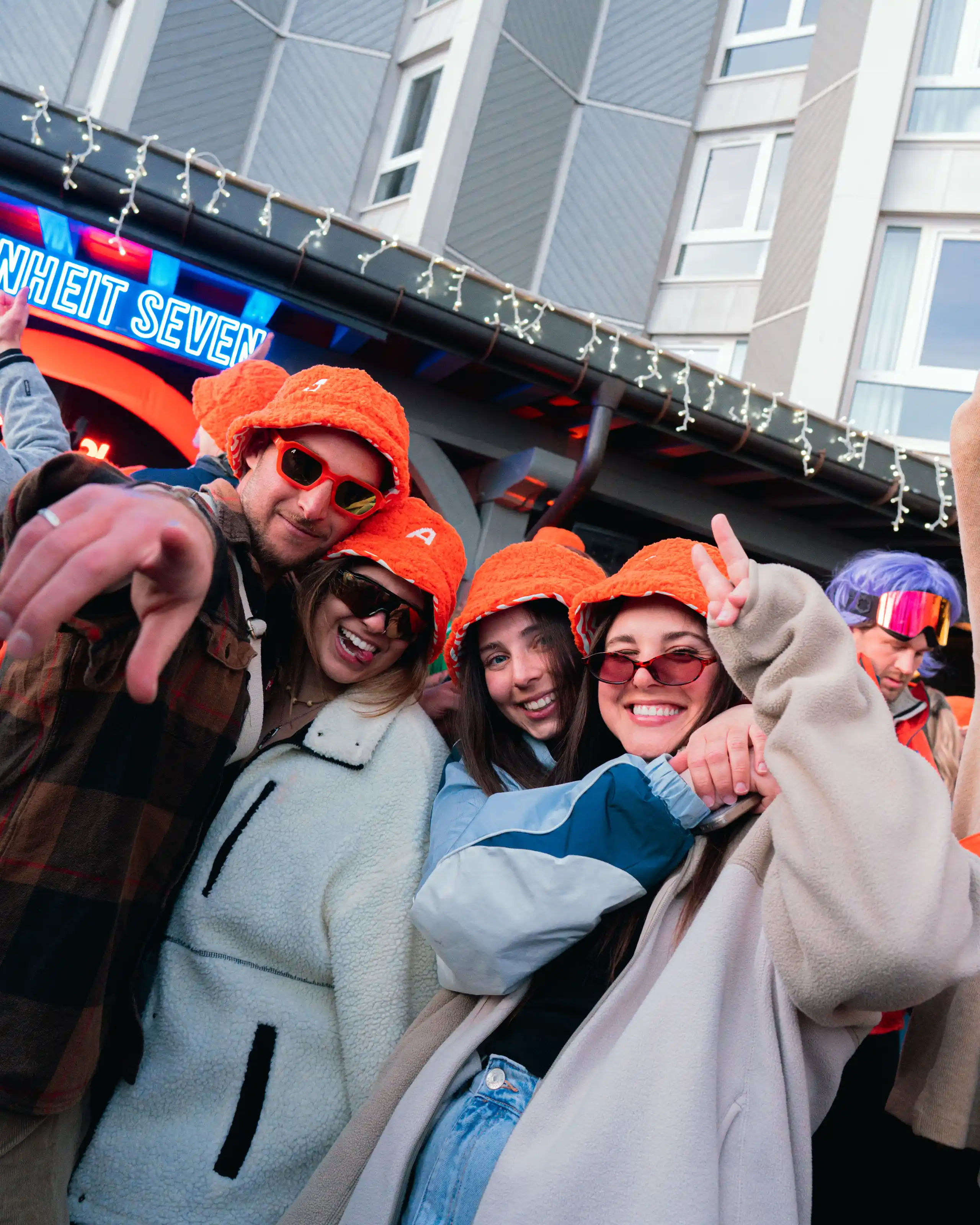 Quatre jeunes souriant, portant des chapeaux orange et des lunettes de soleil, posant ensemble lors d'un événement en extérieur, avec un bâtiment et des guirlandes lumineuses en arrière-plan.