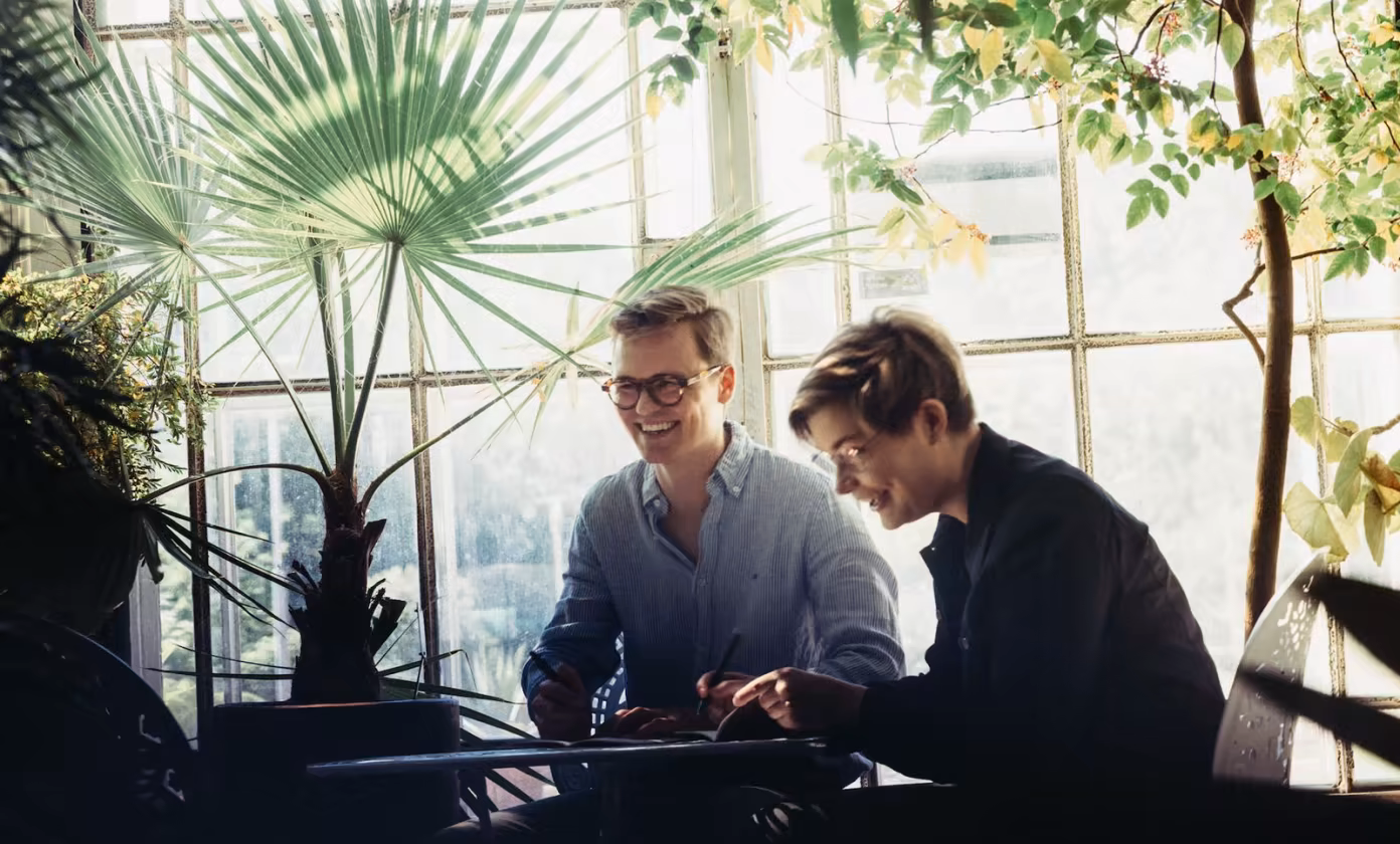 Two people smiling while working together at a table in a bright room with large plants.