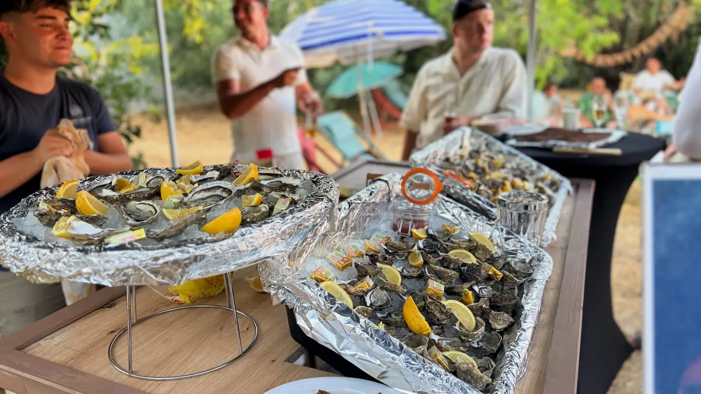 Platters of fresh oysters on ice with lemon wedges at an outdoor event with people in the background.