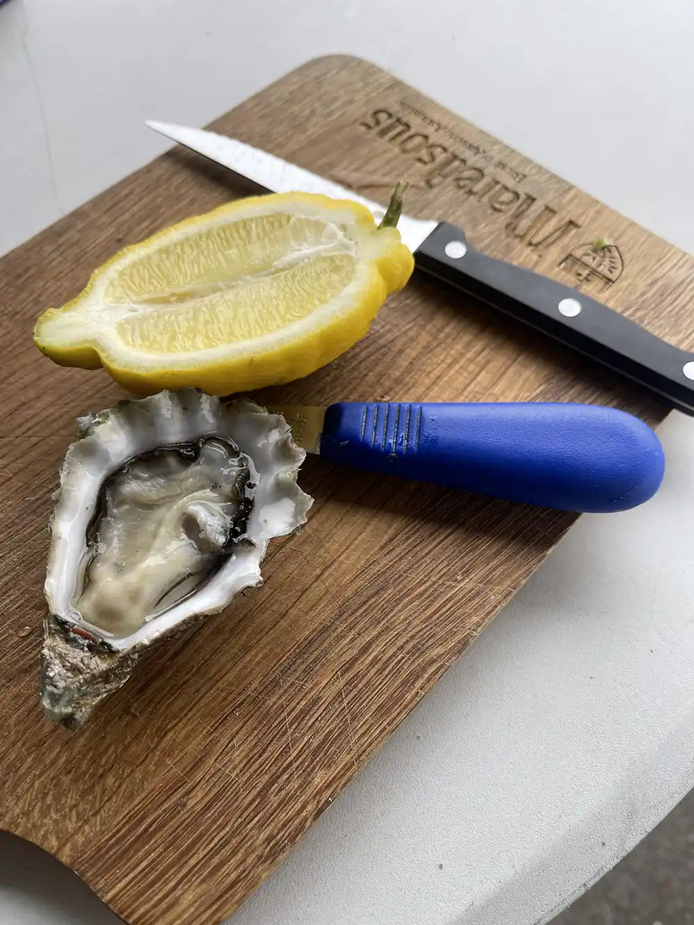Half lemon, opened oyster with blue-handled oyster knife, and a paring knife on a wooden board.
