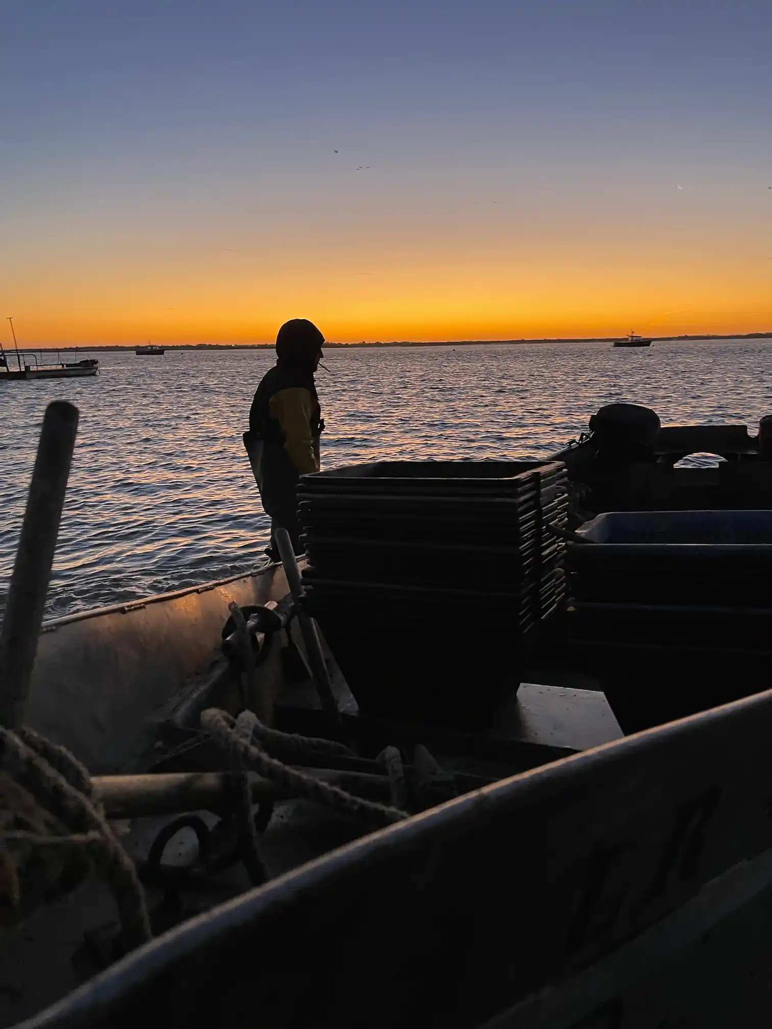 Silhouette of a person on a boat with stacked crates at sunset over calm water with distant boats.