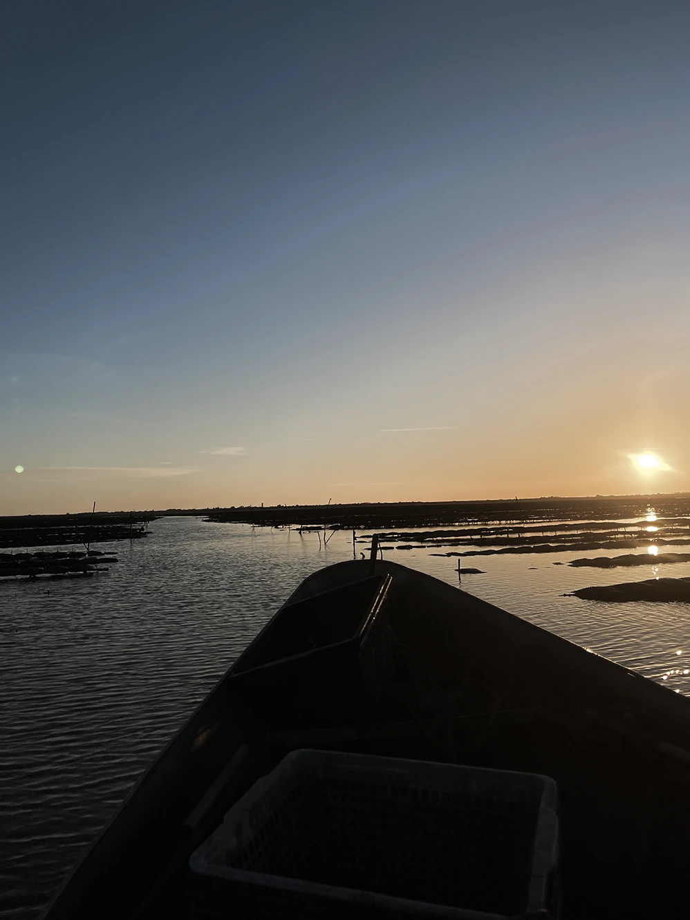 View from a boat on calm water at sunset with clear sky and silhouette of distant shoreline.