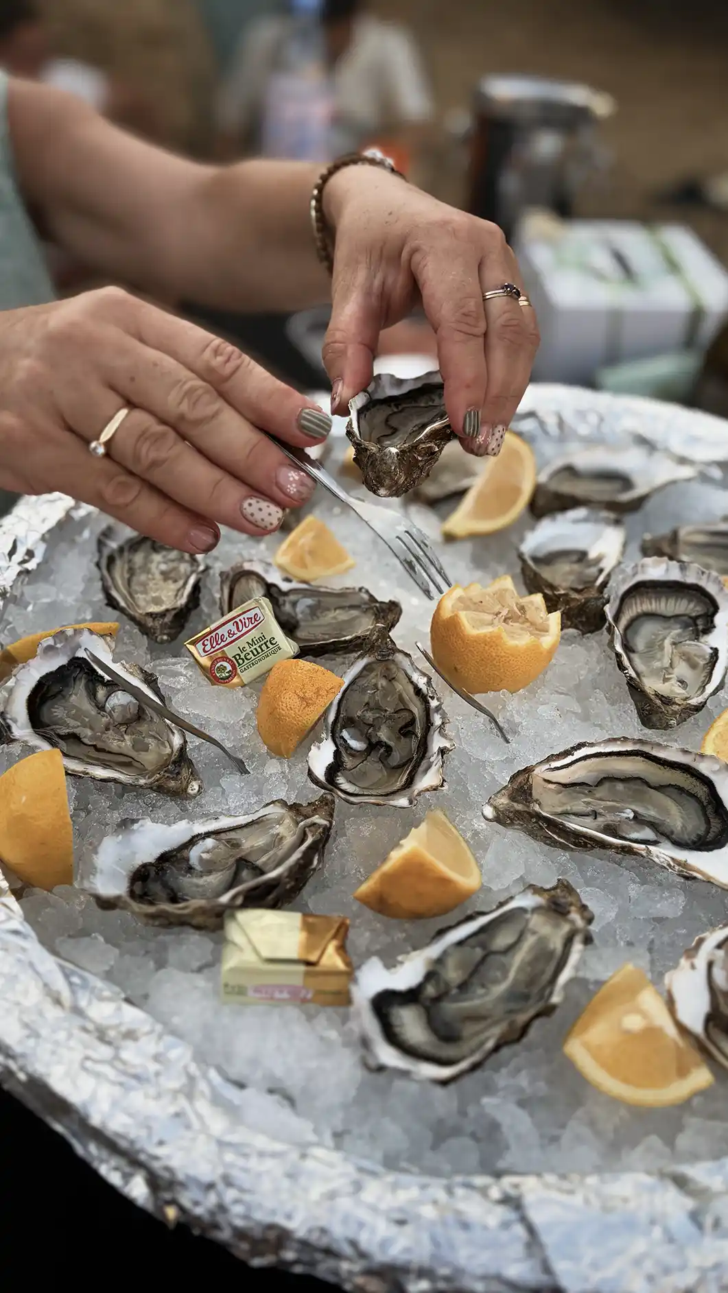 Hands holding an oyster and fork over a tray of oysters on ice with lemon wedges and butter packets.