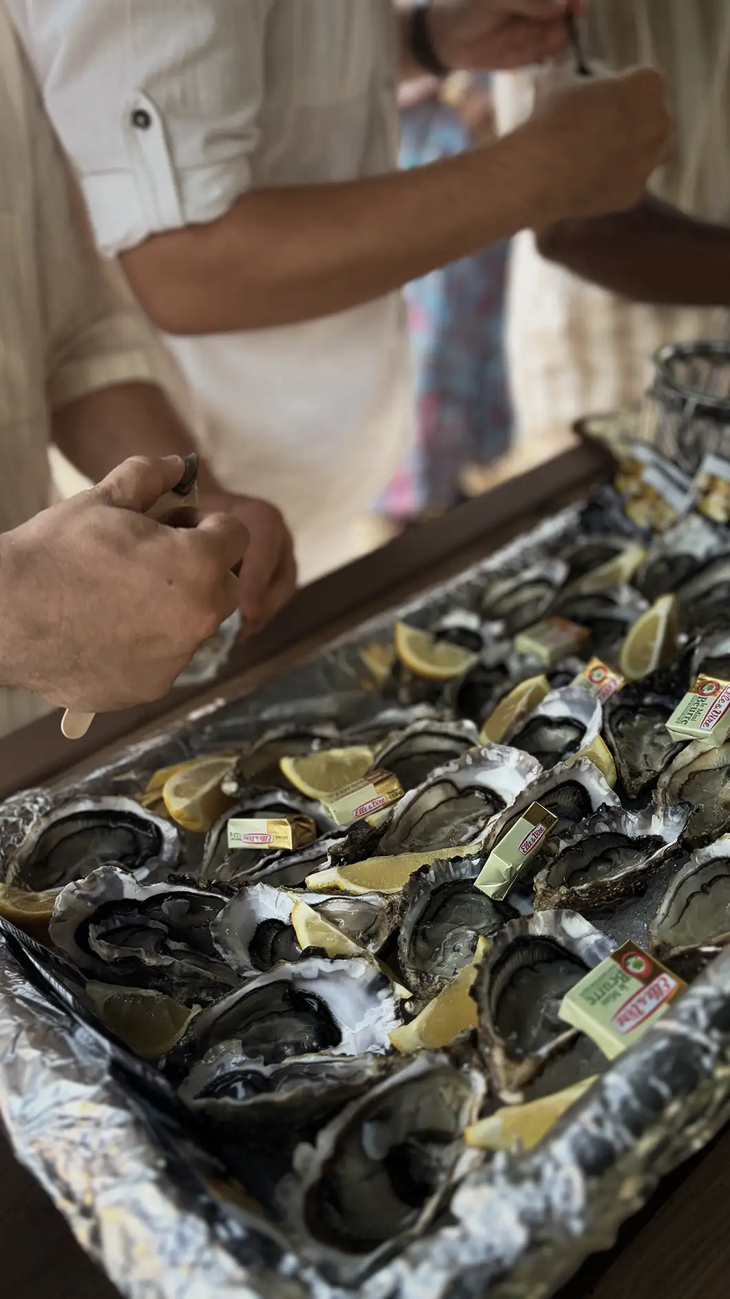 People shucking and eating fresh oysters on a tray with lemon wedges and butter packets.
