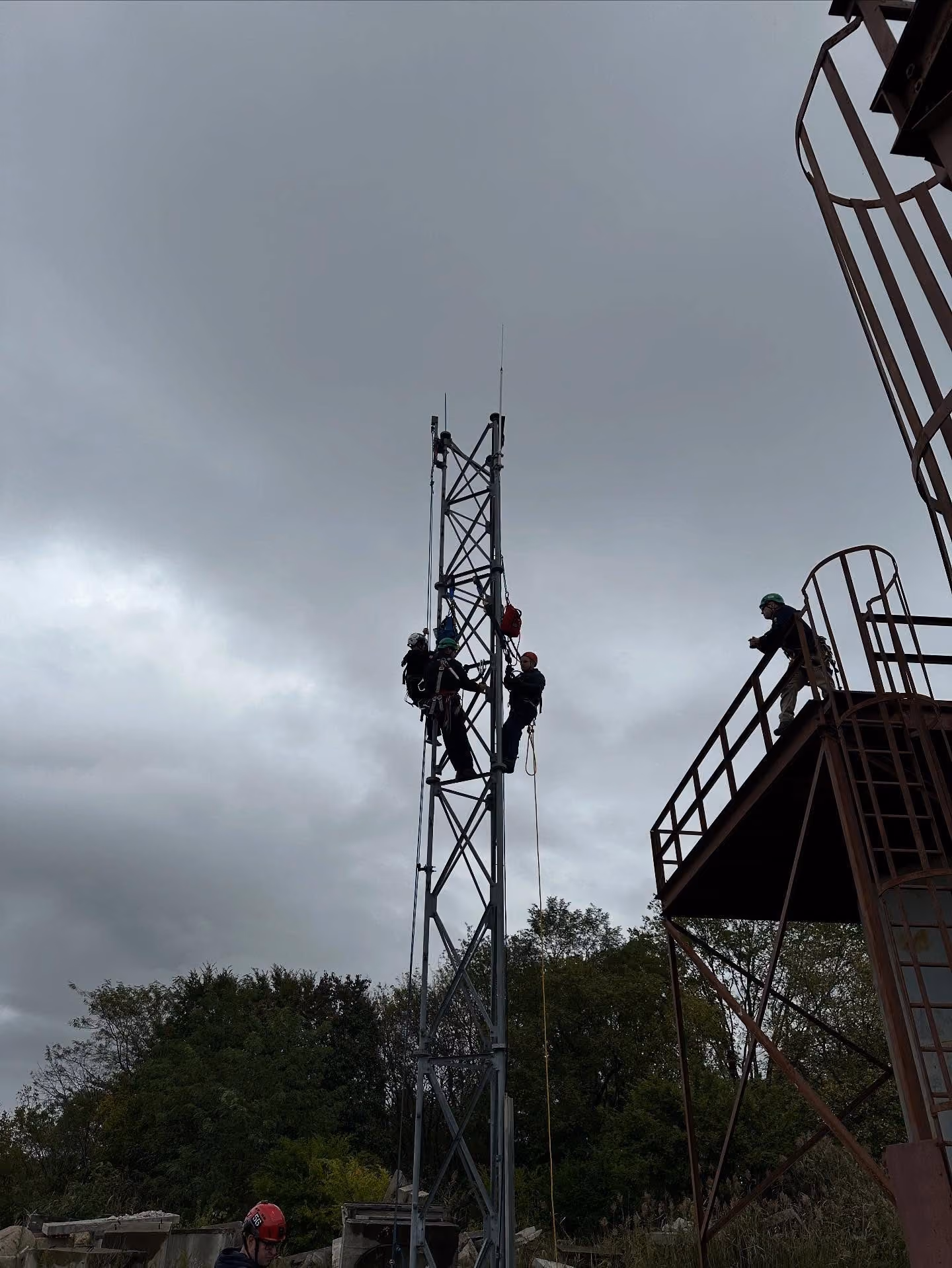 Workers wearing helmets climbing a metal tower for rigging under cloudy sky.