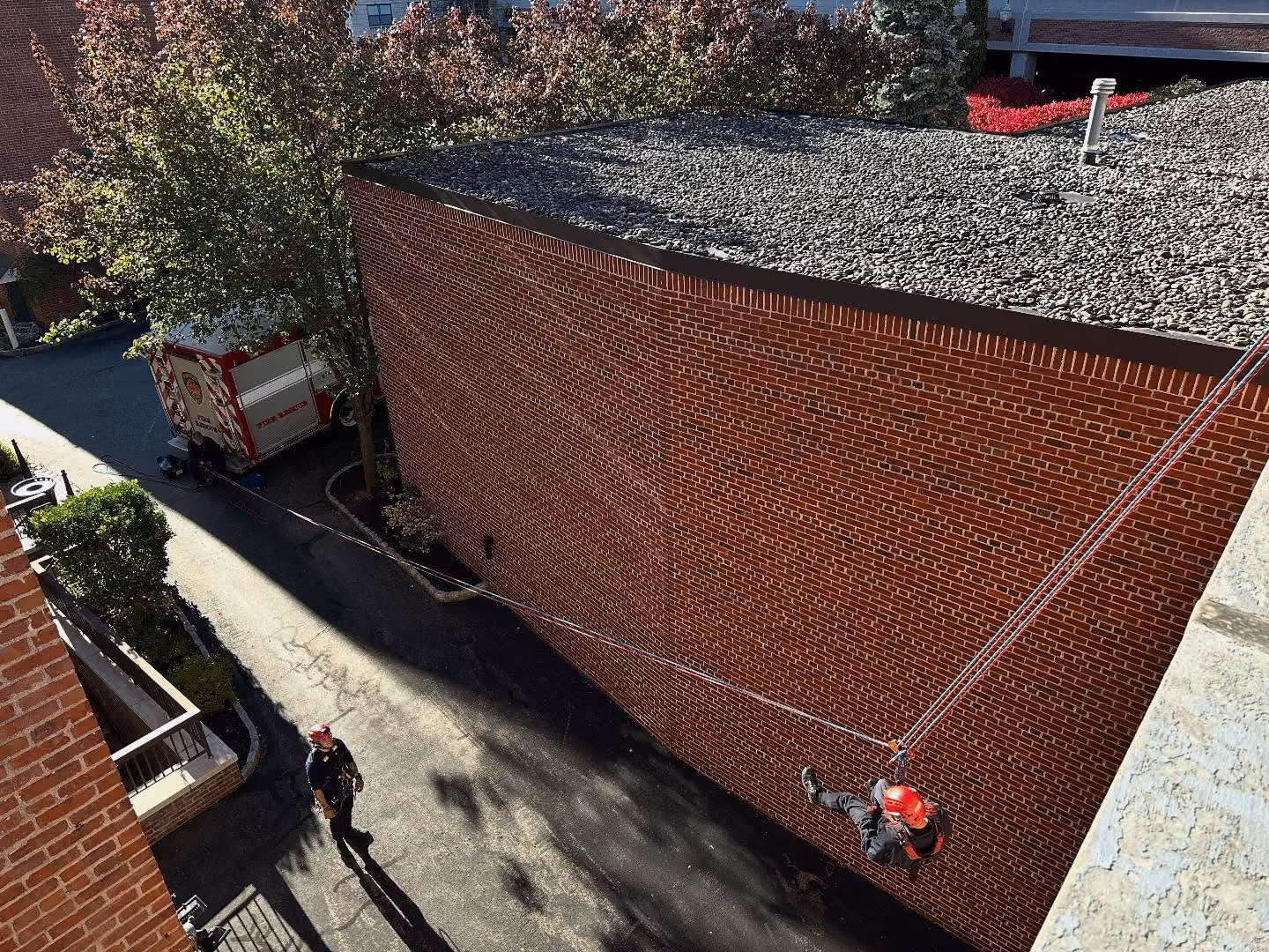 Person rappelling down brick building using ropes and wearing a red helmet with another person below.