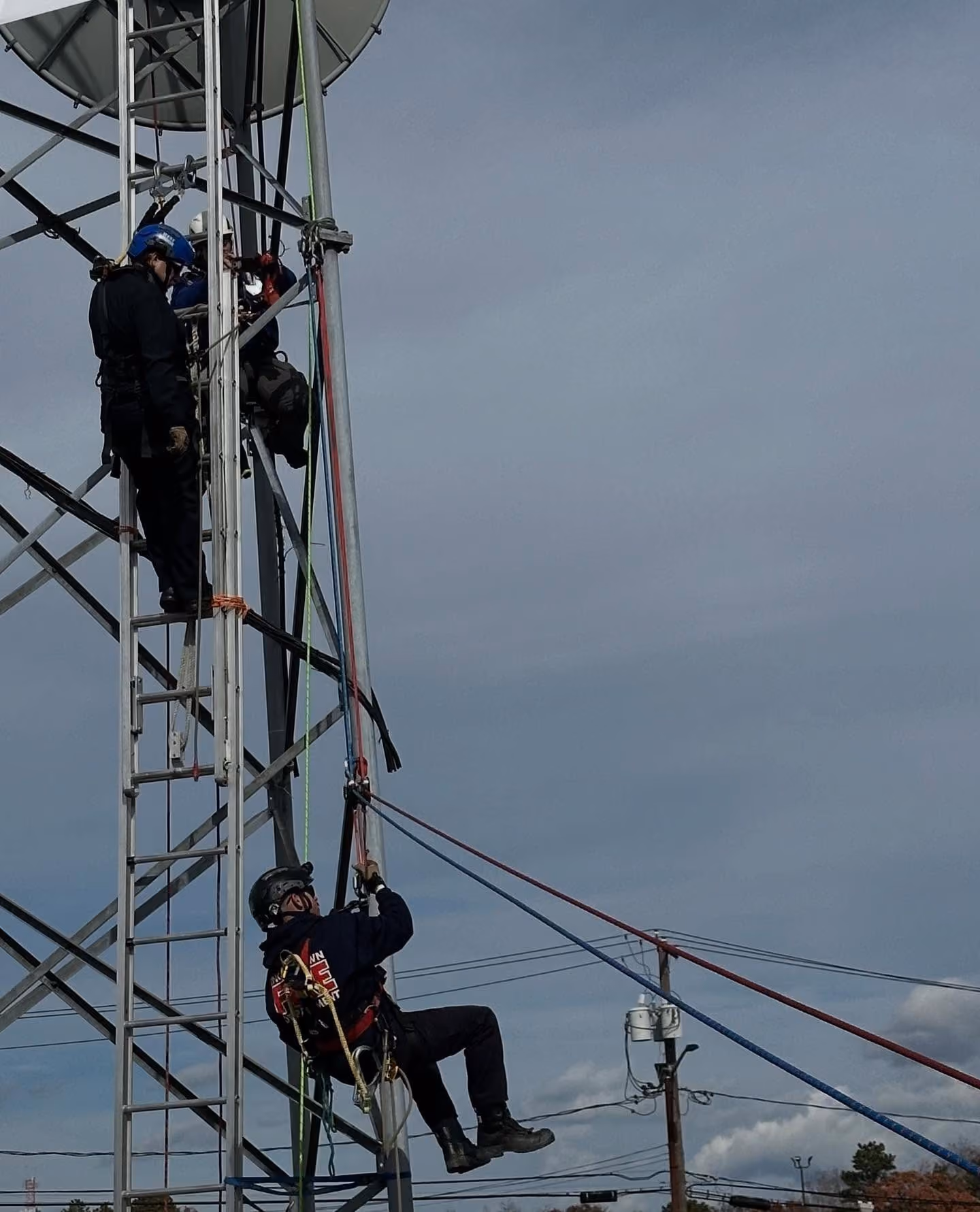 Three workers in helmets climbing and rappelling on a metal tower with ropes on a cloudy day.
