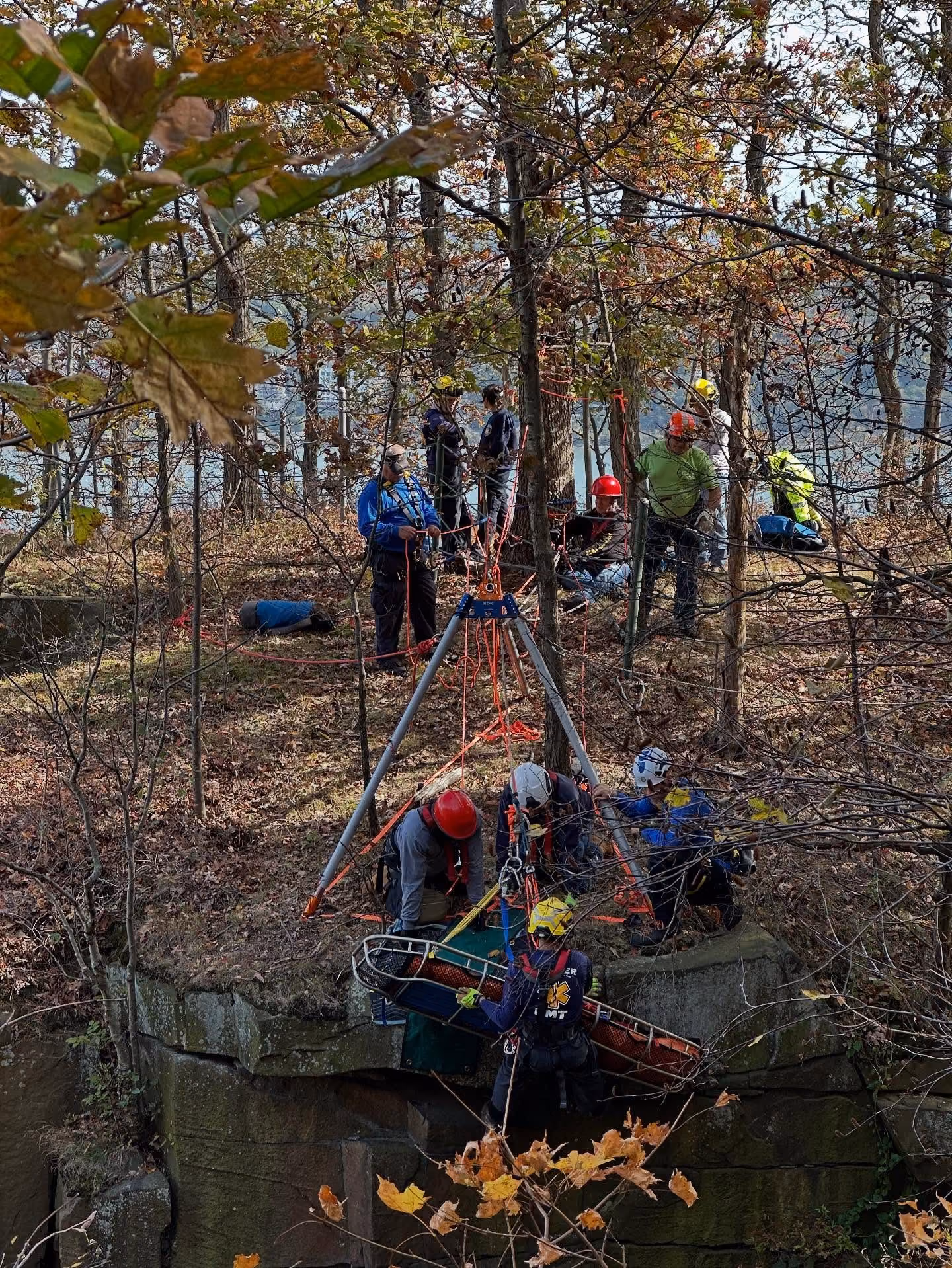 Rescue team using ropes and a tripod to lift a stretcher near a rocky cliff in a forest.
