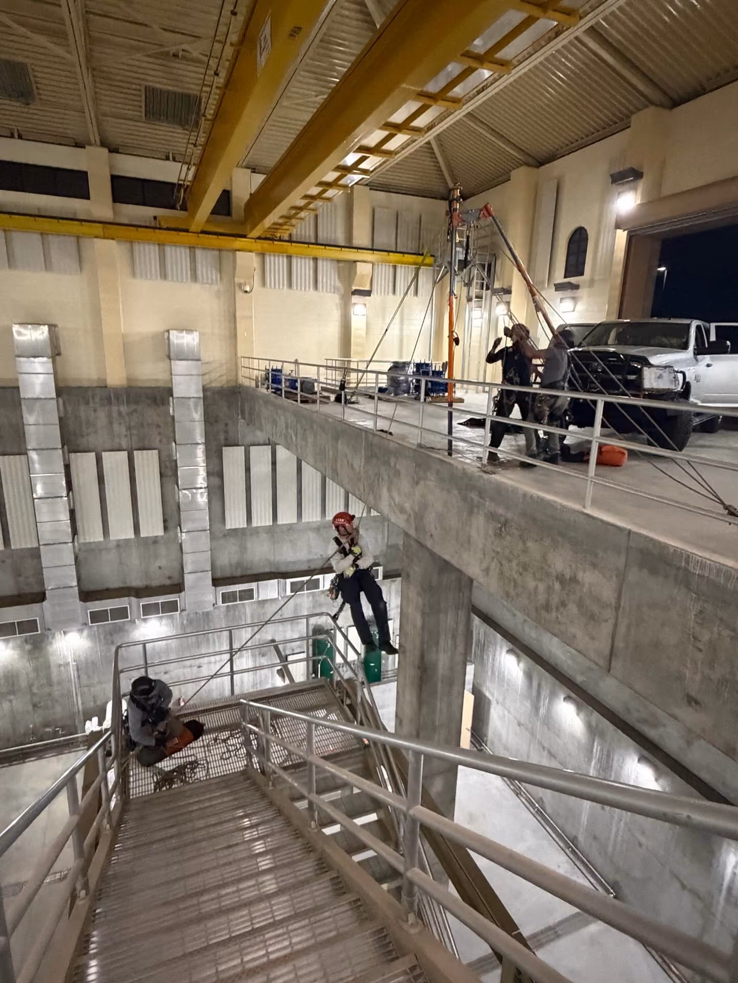 Workers using safety harnesses rigging ropes inside an industrial building near stairs.