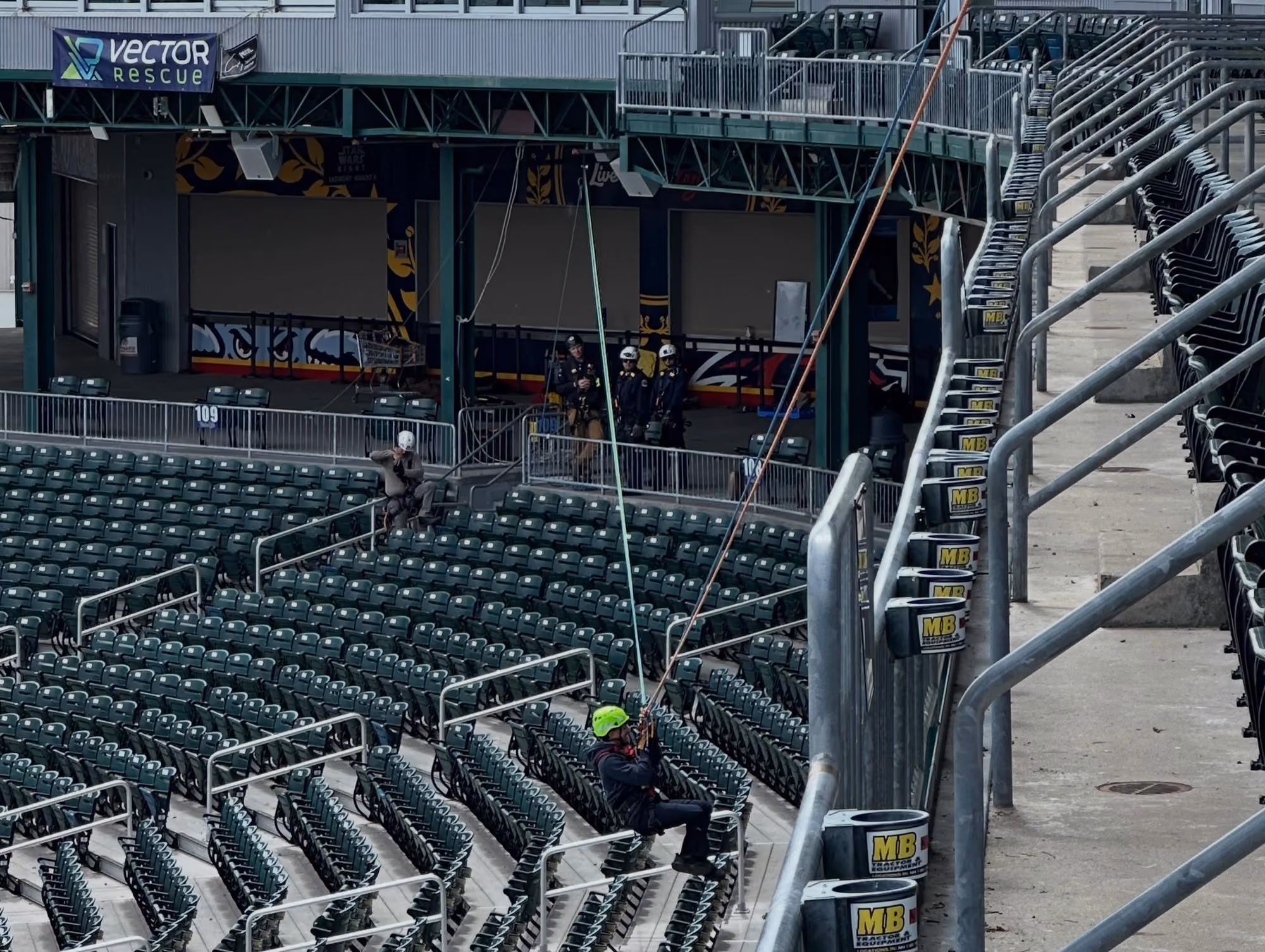 Person in safety gear suspended by ropes above stadium seats with two others observing nearby.