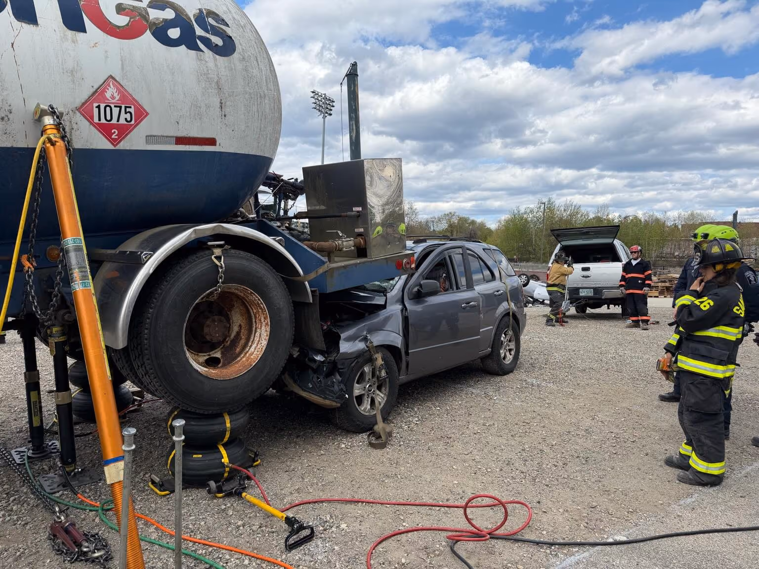 Firefighters conduct rescue training with crushed car under a gas tanker truck on gravel lot.