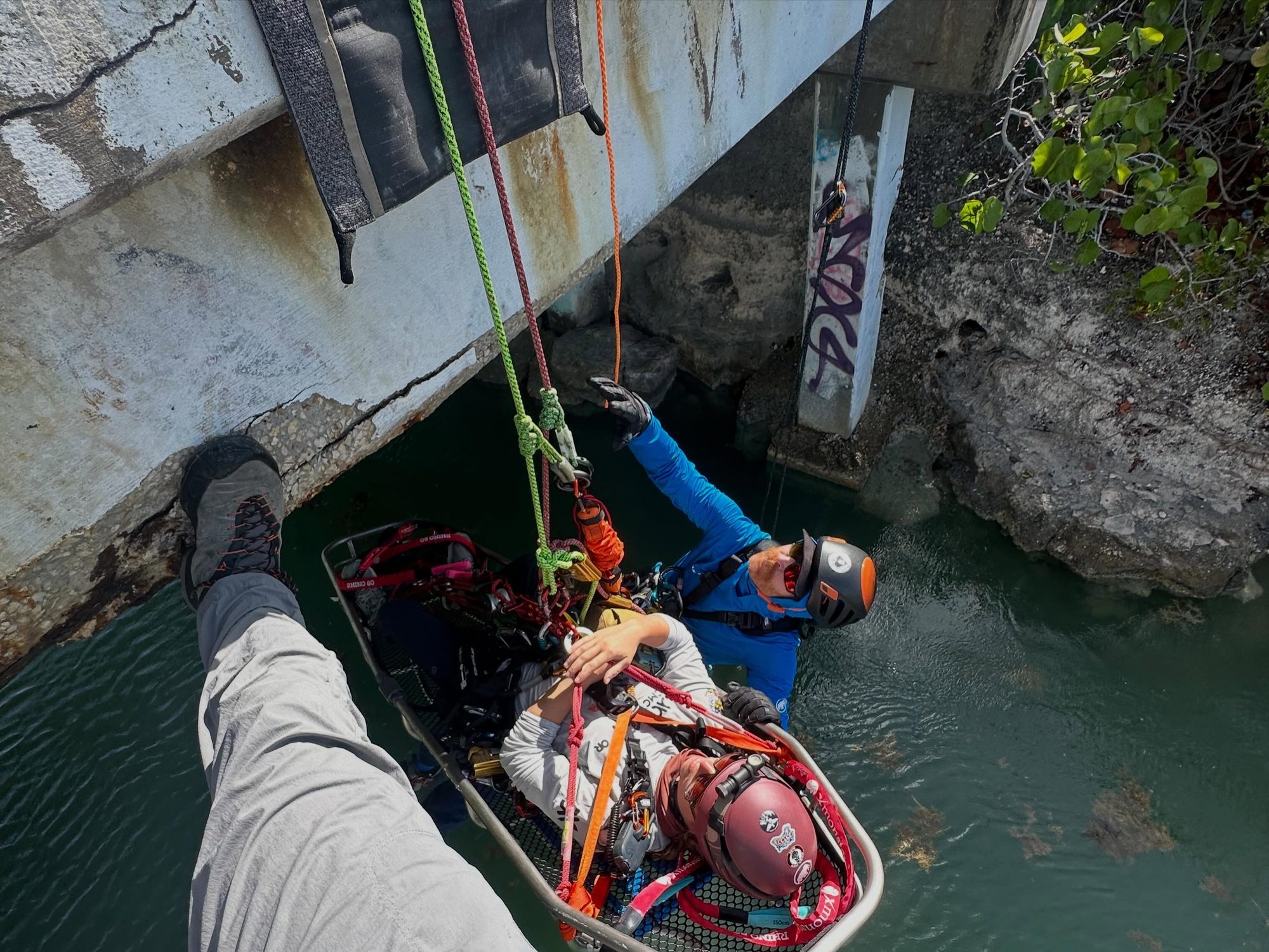 Rescuer lowering a person on a stretcher with ropes under a concrete bridge over water.