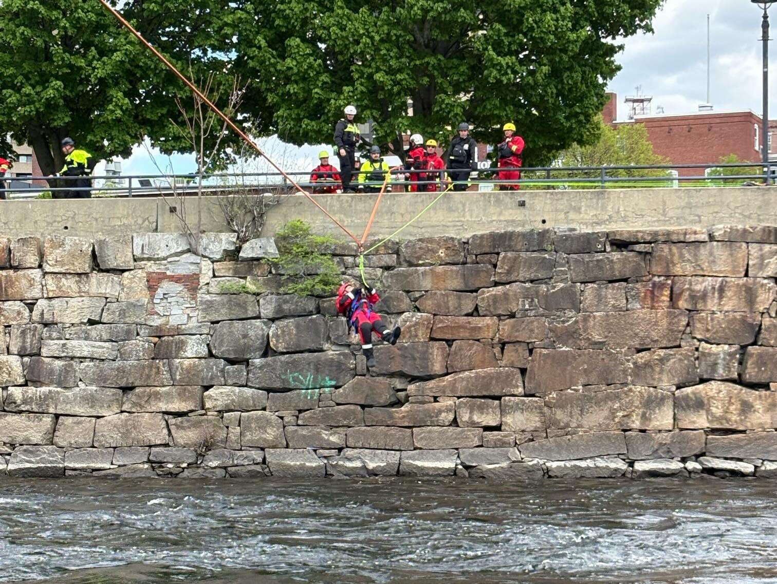 Person in rescue gear suspended by ropes from a stone wall over flowing water with several people watching above.
