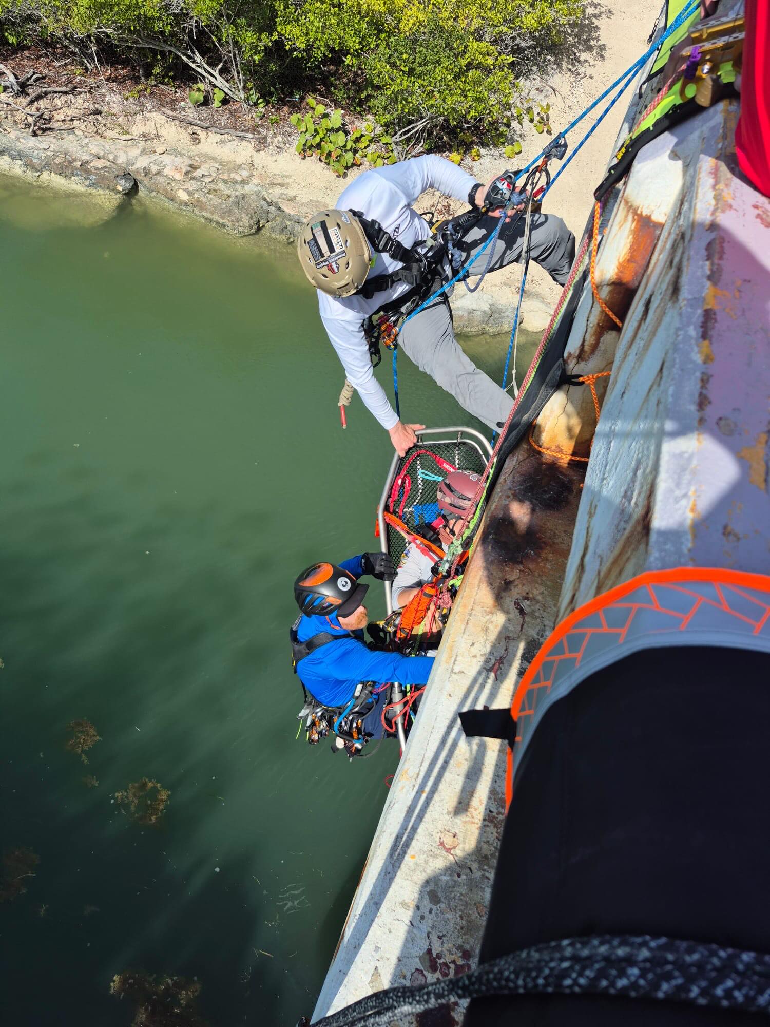 Two rescue workers with helmets and harnesses lowering a person in a stretcher over green water.