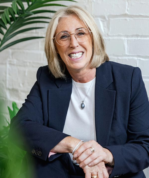 Dr Sarah Carlick smiling as she sits for a photograph against a white brick wall.