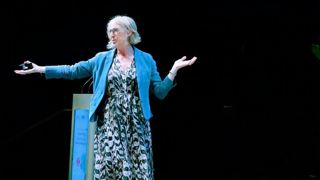Dr Sarah Carlick speaks on stage with arms extended, wearing patterned dress and blue jacket.