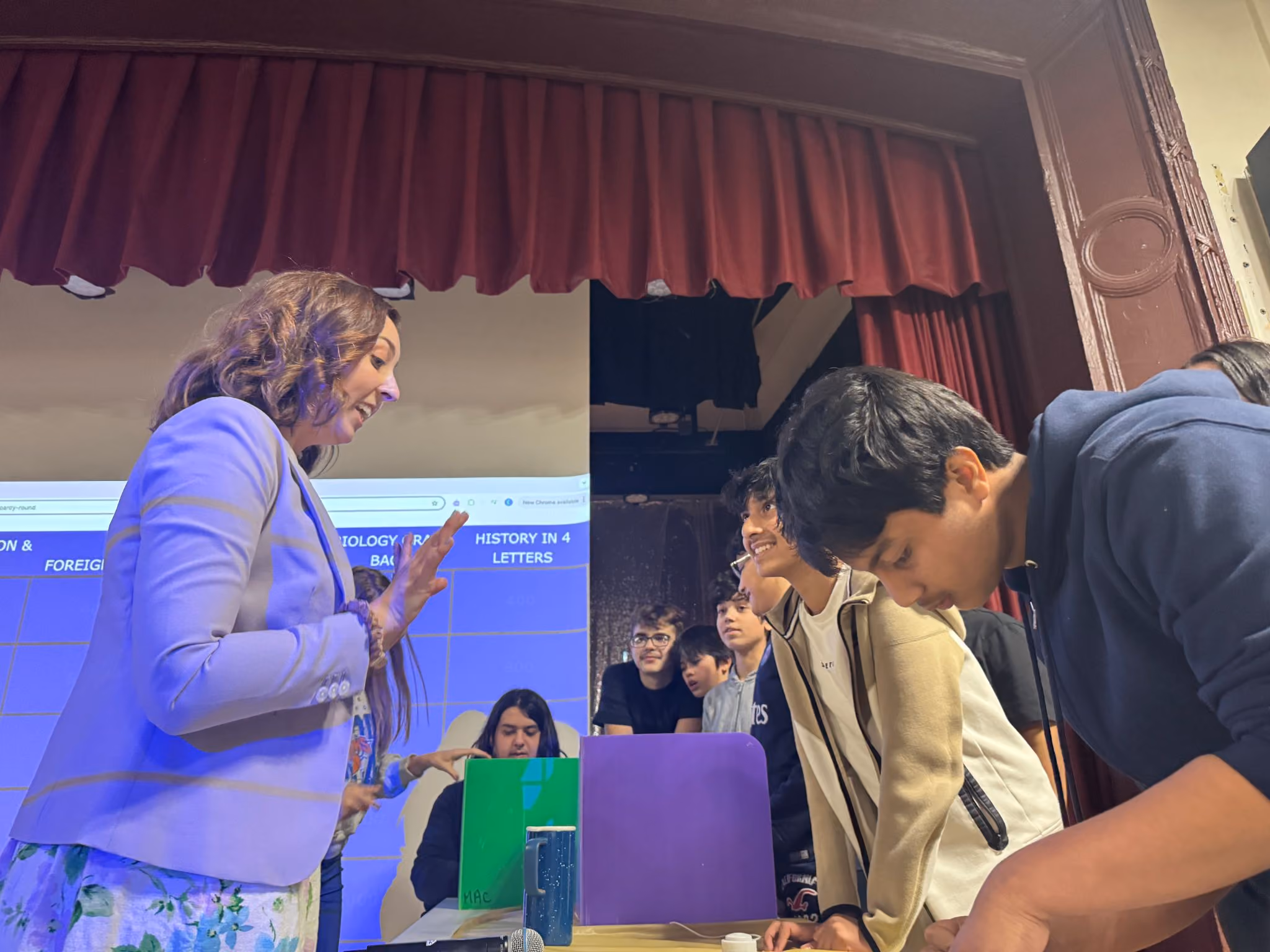 Teacher hosting MAA and RBG Jeopardy as student teams answer on stage