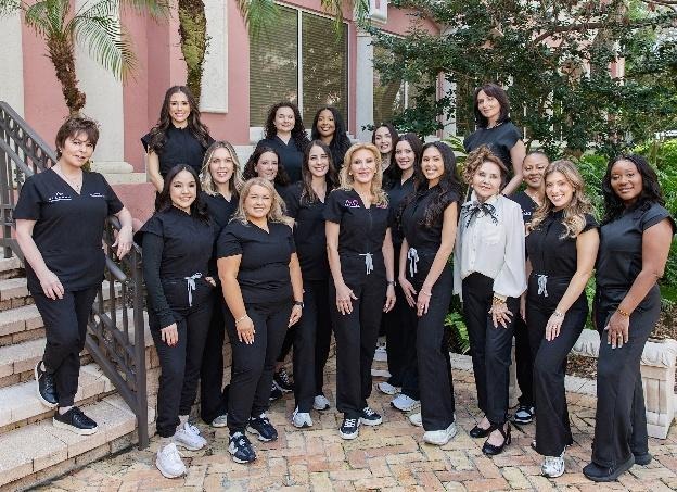 The image shows a group of dental professionals standing together, dressed in white coats, with some wearing black bowties, positioned in front of a building with palm trees and a staircase.AI-generated content may be incorrect.