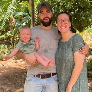 Smiling couple outdoors holding a baby in greenery with trees and plants in the background.