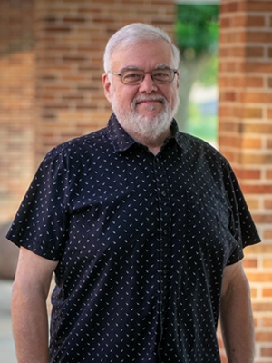 Smiling older man with white hair, beard, and glasses in a black patterned shirt outdoors by brick pillars.