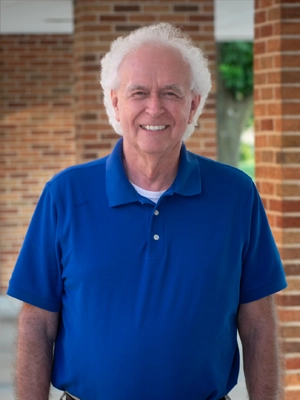 Smiling elderly man with white hair wearing a blue polo shirt standing outdoors near brick columns.
