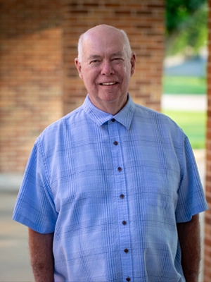 Smiling older man in a light blue plaid short-sleeve shirt standing outdoors by a brick wall.