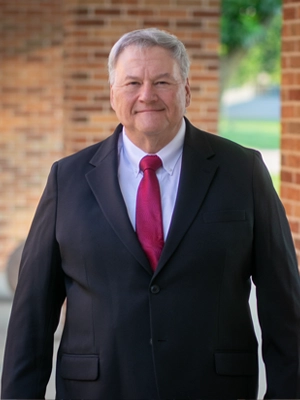 Smiling man wearing a black suit and red tie standing outdoors near a brick wall.