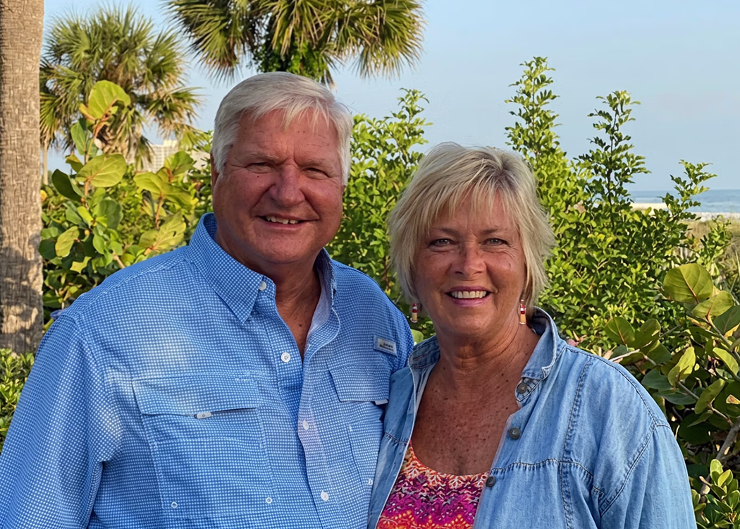Smiling older man in blue shirt and woman in patterned top and denim shirt stand outdoors by greenery.