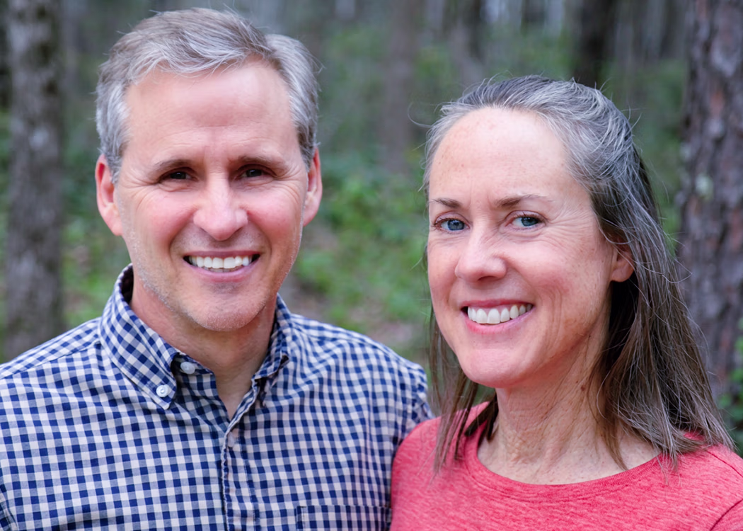 Smiling middle-aged man in checkered shirt and woman in red top outdoors with trees in background.