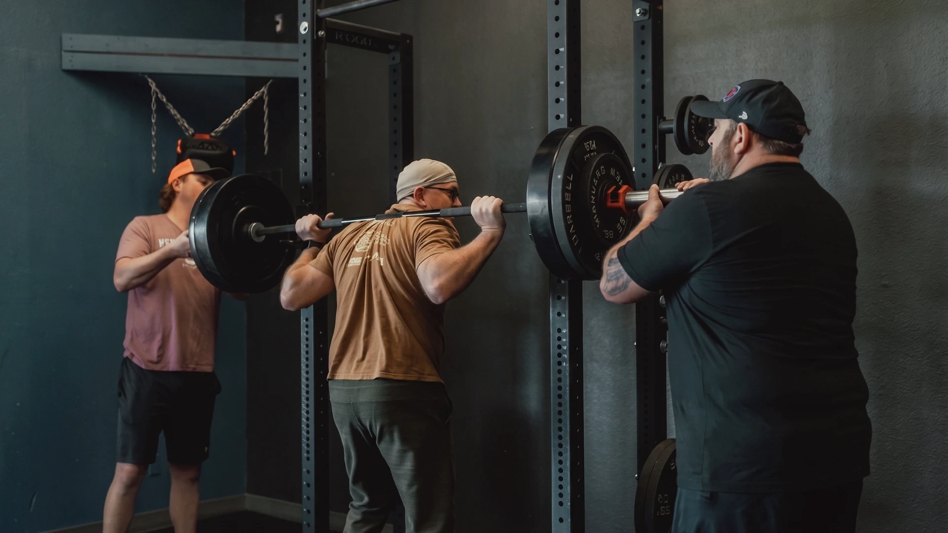 A coach and athlete working together during a barbell squat in a strength training gym, with a spotter assisting on either side of the loaded barbell.