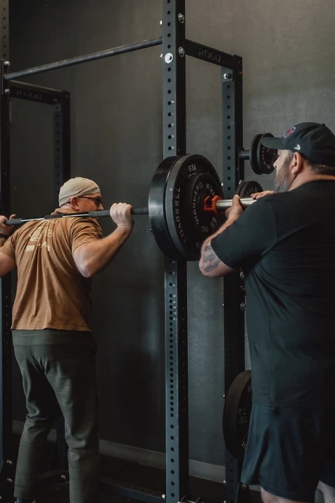 A coach and athlete working together during a barbell squat in a strength training gym, with a spotter assisting on either side of the loaded barbell.
