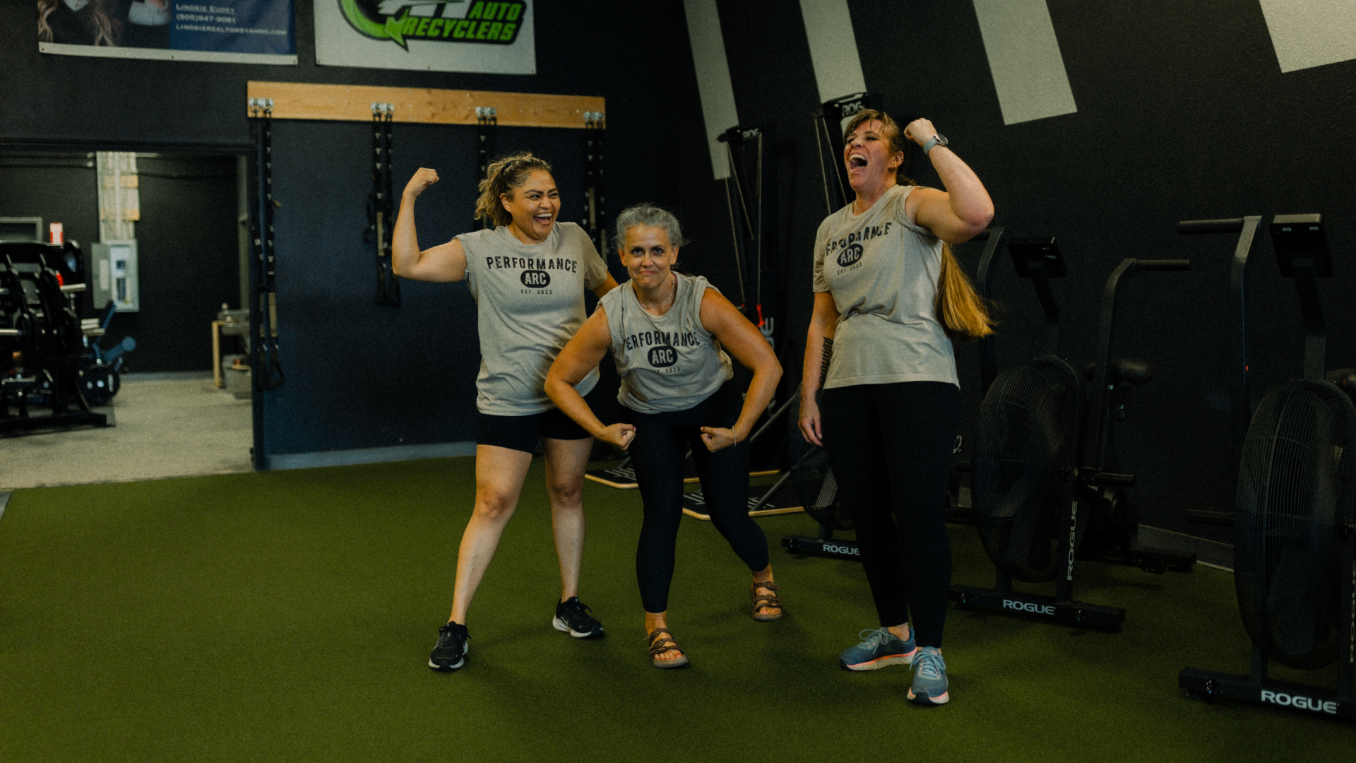 Three women in matching Performance ARC shirts pose playfully and flex their arms together on indoor turf at a gym, with exercise equipment in the background.