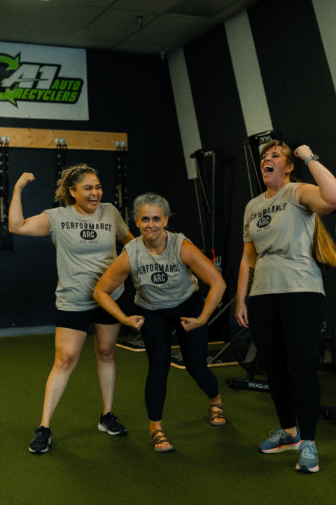 Three women in matching Performance ARC shirts pose playfully and flex their arms together on indoor turf at a gym, with exercise equipment in the background.