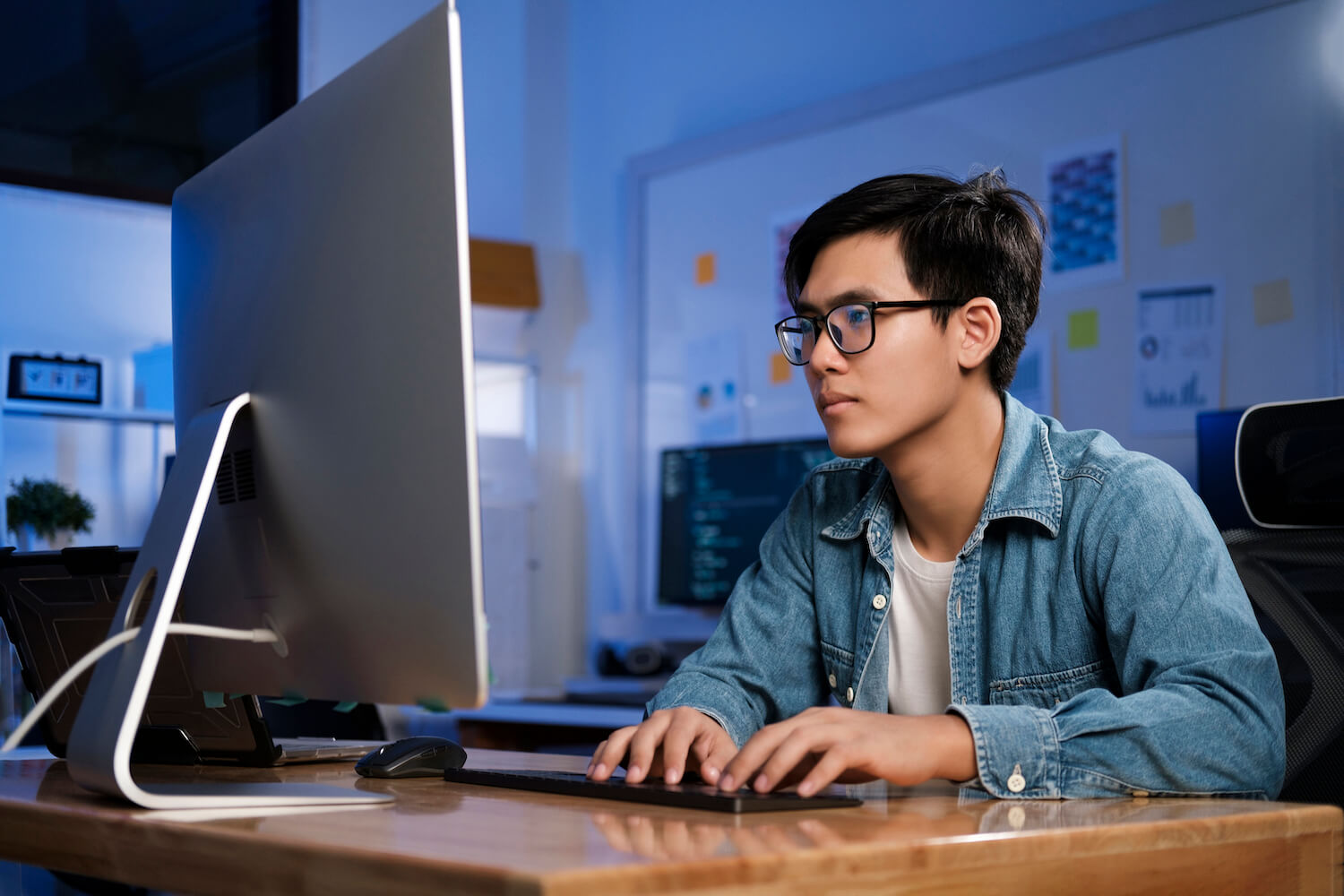 Young man in glasses and denim jacket focused on coding at desktop computer in dimly lit office.
