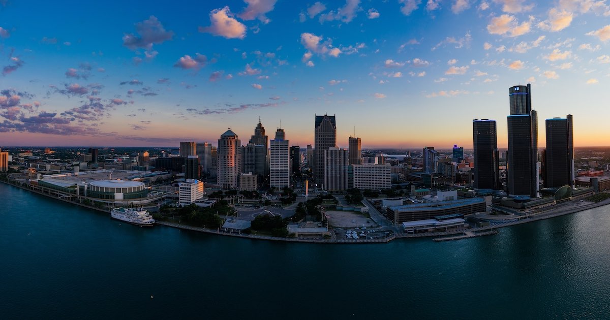 Panoramic view of a city skyline at sunset with tall buildings along a riverfront under a partly cloudy blue sky.